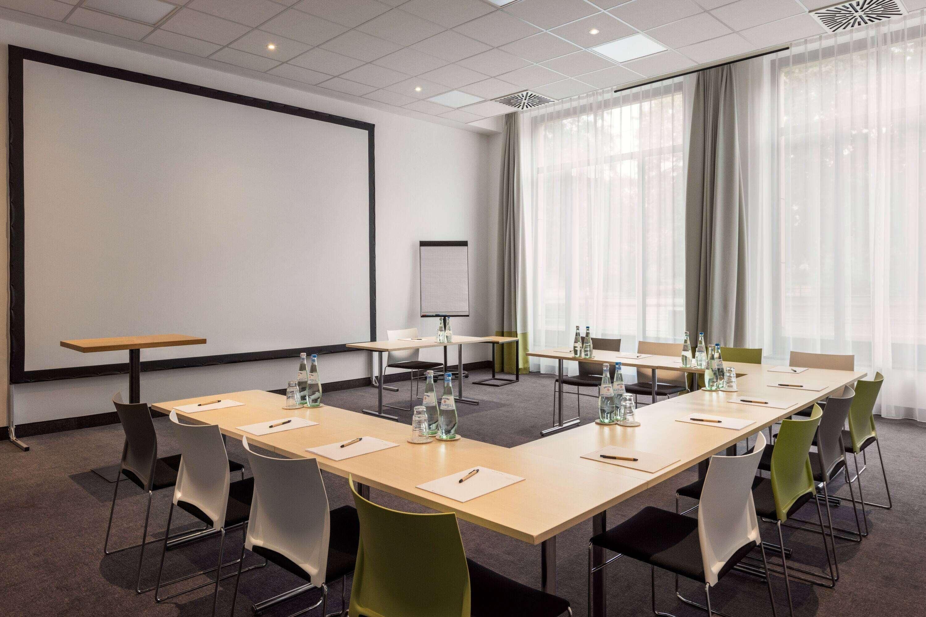 A conference room with a large screen, projector, and a whiteboard. Tables are set with chairs, water bottles, and notepads.