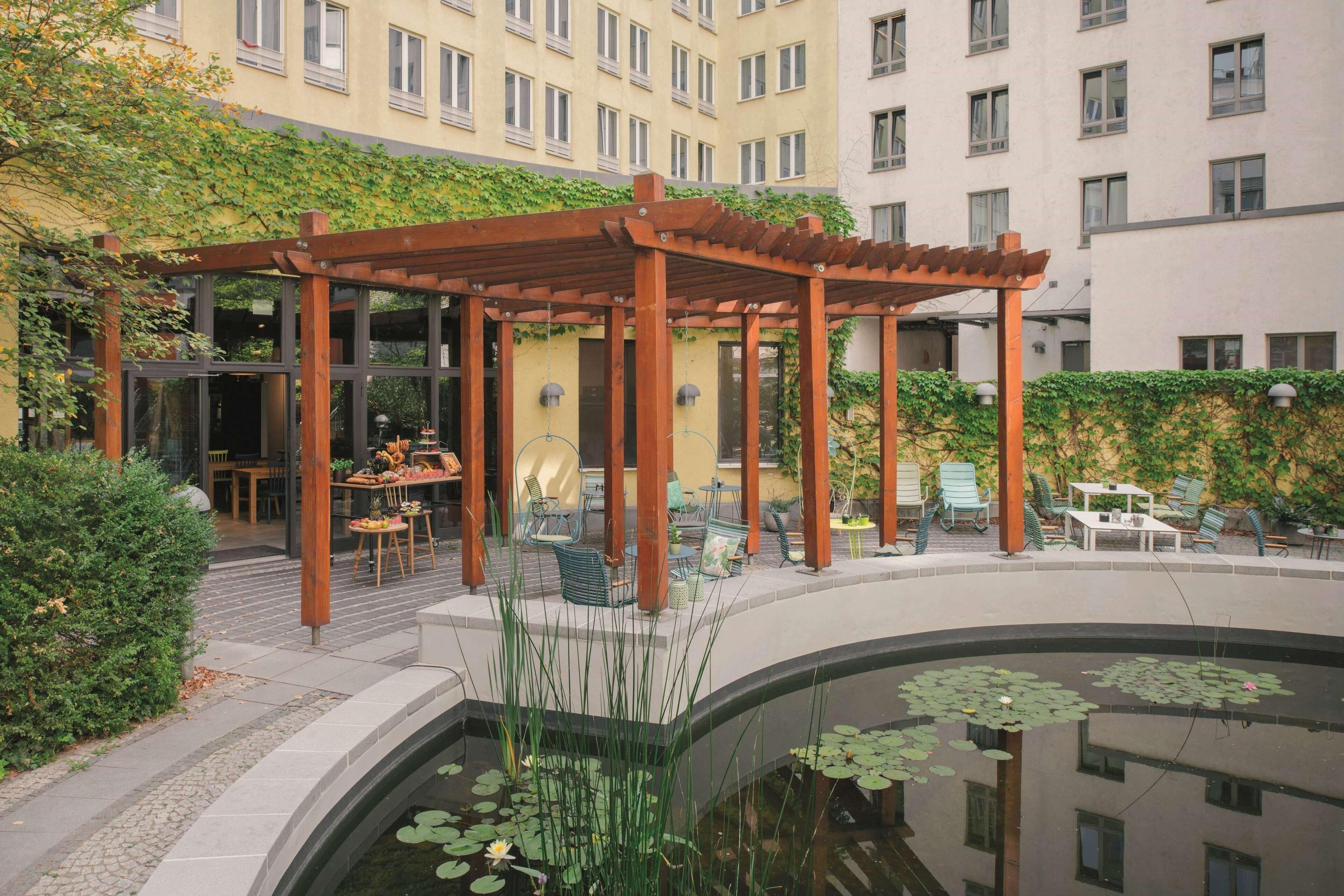 Un patio con una pérgola de madera, mobiliario exterior, un pequeño estanque con nenúfares y un edificio con múltiples ventanas al fondo.