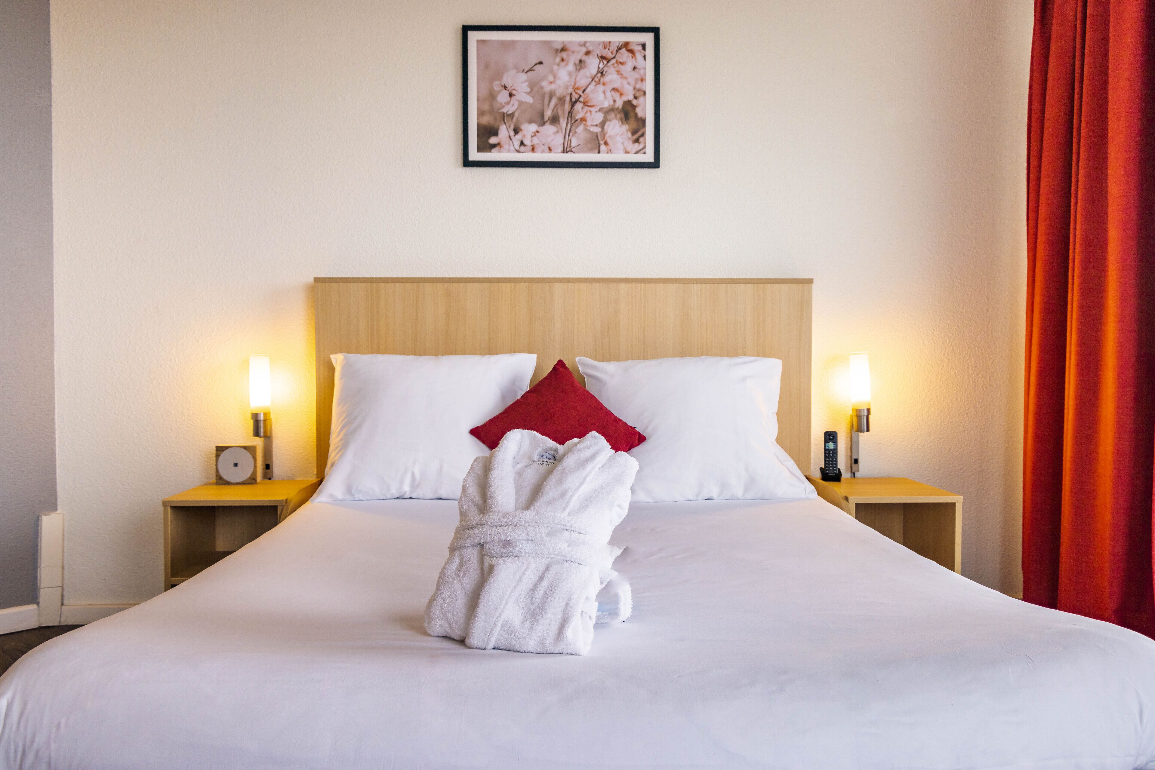 Een keurig opgemaakt bed met witte lakens, een rood kussen en een witte badjas. Twee nachtkastjes met lampen en een ingelijst beeld aan de muur boven het bed.