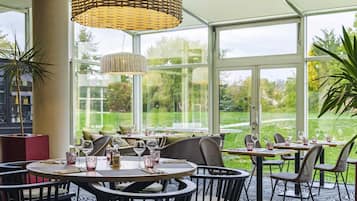 Un restaurante con mesas redondas, sillas de mimbre y amplios ventanales que dan a un jardín.