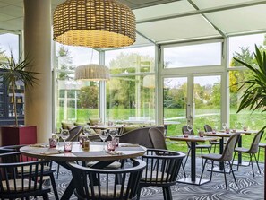 Un restaurante con mesas redondas, sillas de mimbre y amplios ventanales que dan a un jardín.