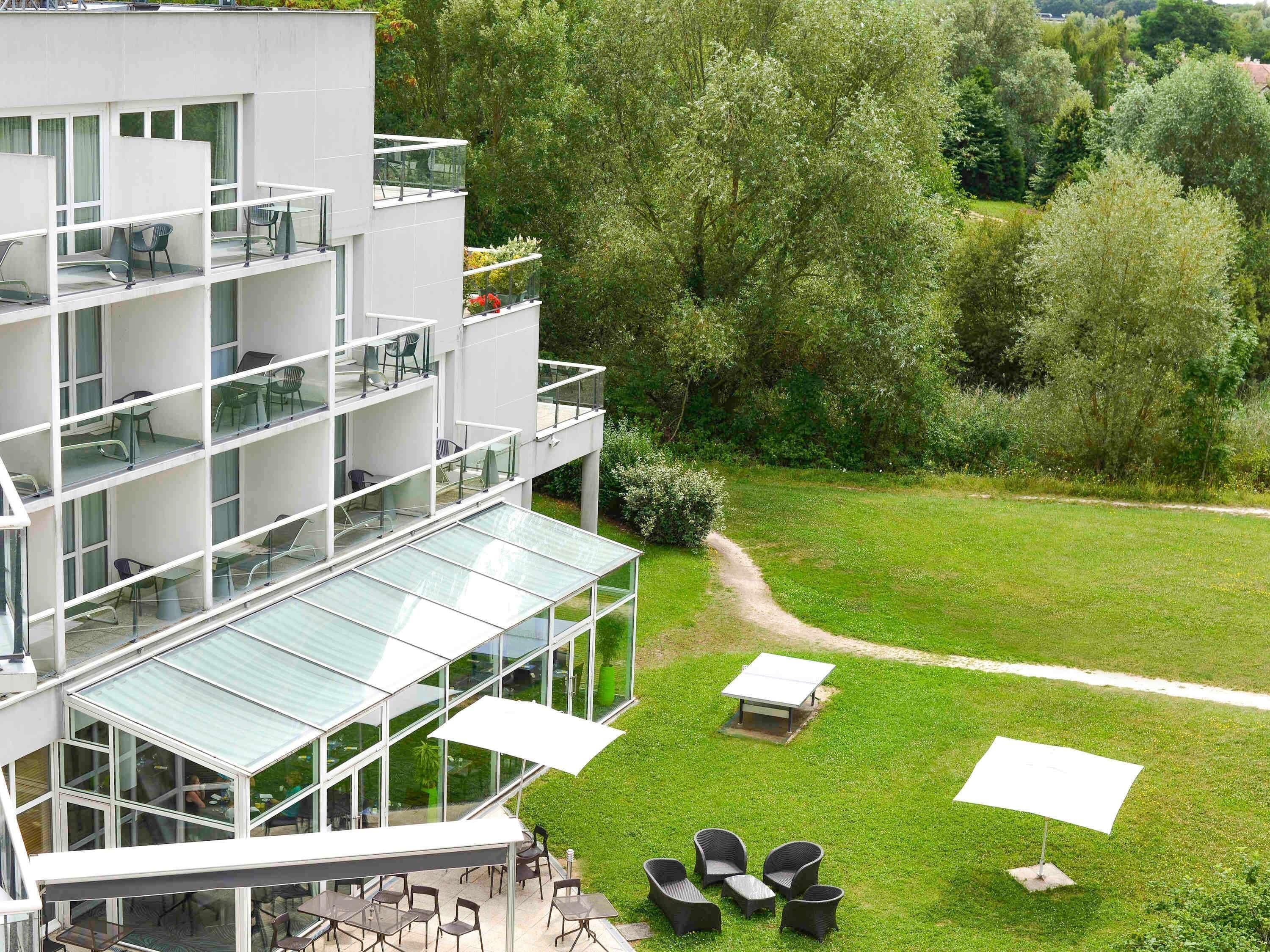 Un bâtiment moderne à plusieurs étages, doté d’une terrasse et d’une véranda en verre.. Hôtel Novotel Senart Golf de Greenparc
