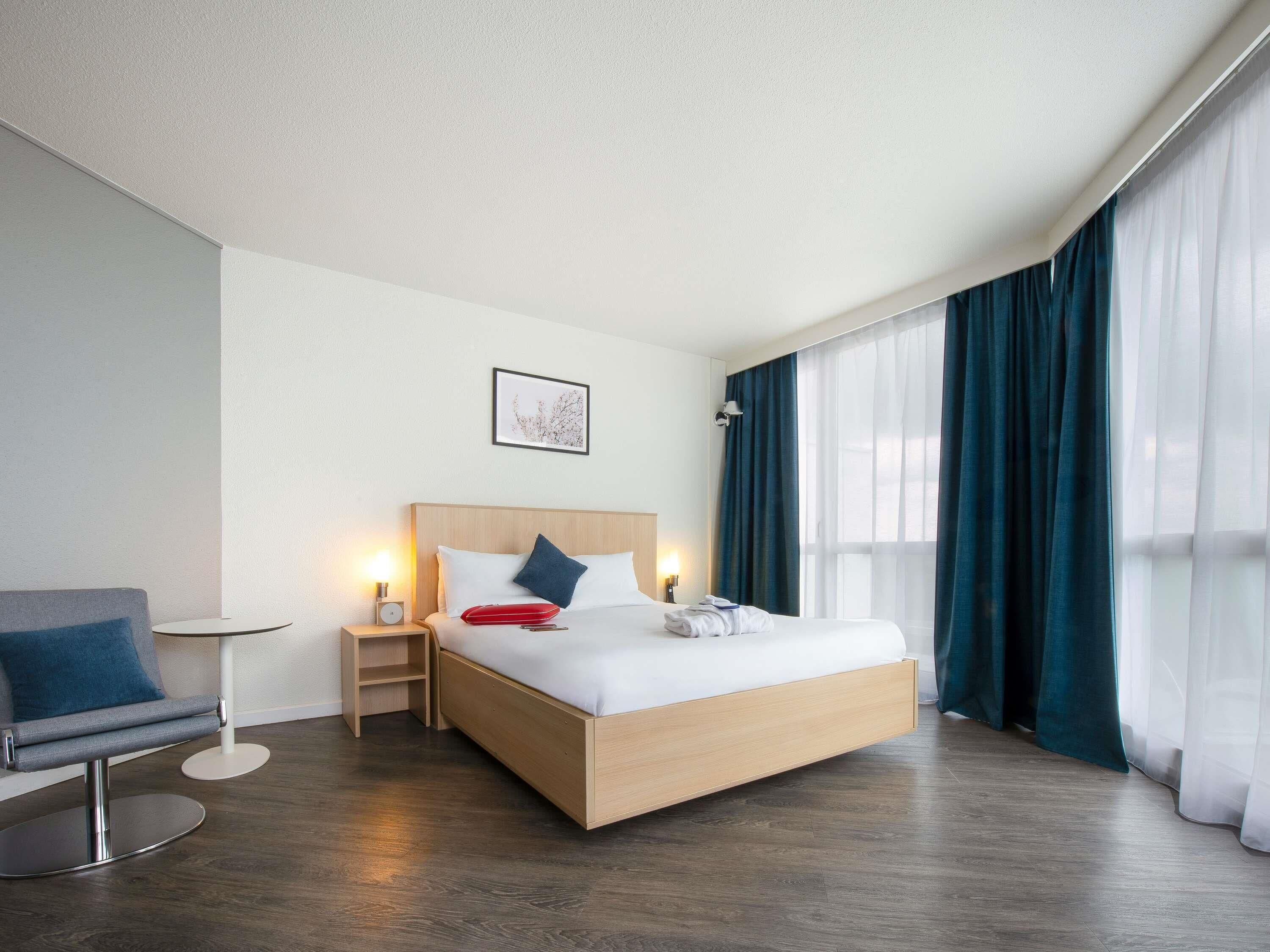 Une chambre d’hôtel moderne avec un lit en bois, une table de chevet, une chaise et une grande fenêtre avec des rideaux.