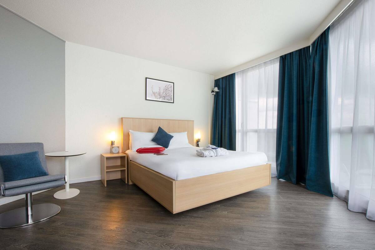 A modern hotel room with a wooden bed, a bedside table, a chair, and a large window with curtains.
