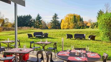 An outdoor dining area with tables and chairs, surrounded by a grassy field and trees.