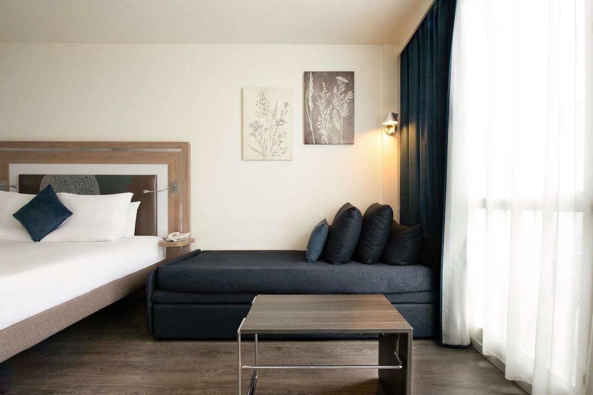 A hotel room with a bed, a sofa, a small table, and a wall-mounted lamp.