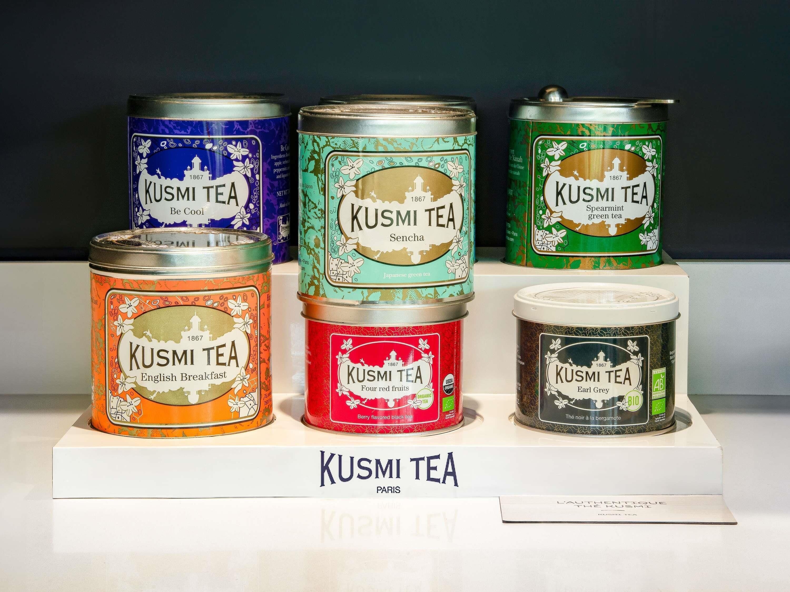 Six cans of Kusmi Tea in various colors, each with a distinct label, displayed on a white stand.