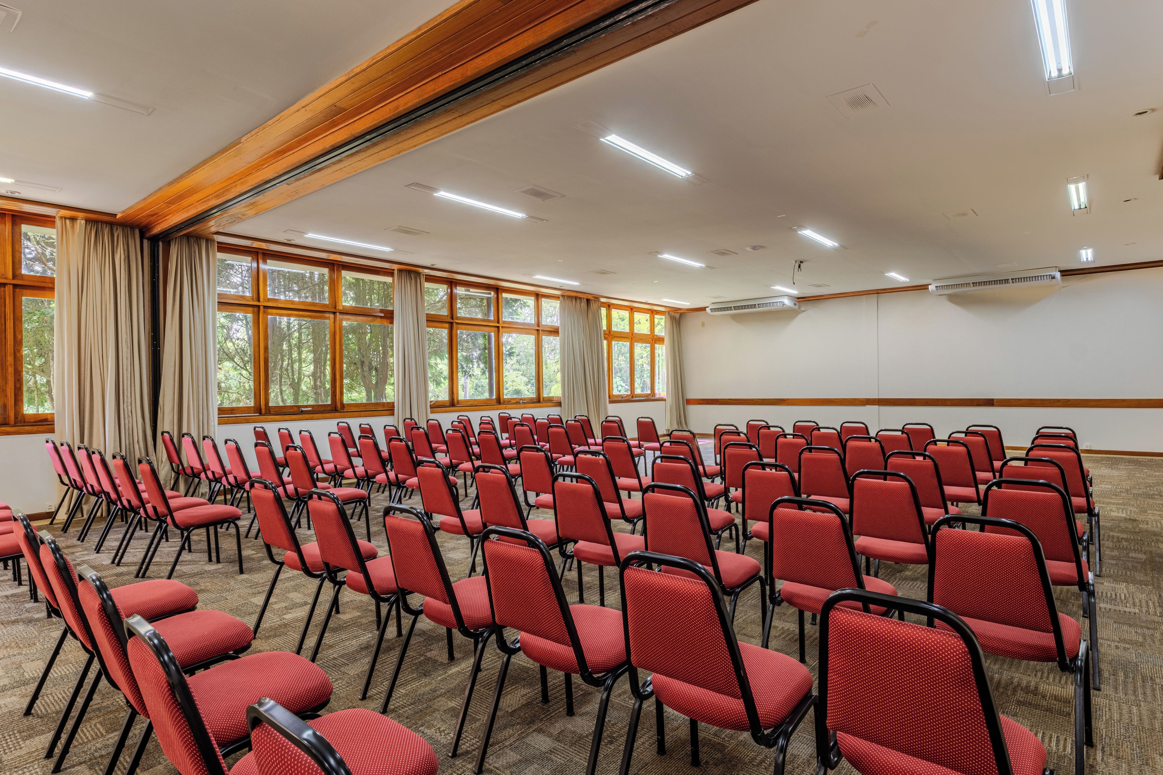 Uma sala de conferência com fileiras de cadeiras vermelhas, janelas amplas com cortinas e iluminação no teto.