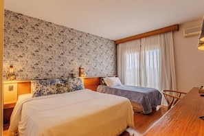 A hotel room with two beds, a wooden desk, a nightstand, and a wall with a floral pattern. - Serra Alta Hotel (Sao Bento do Sul)