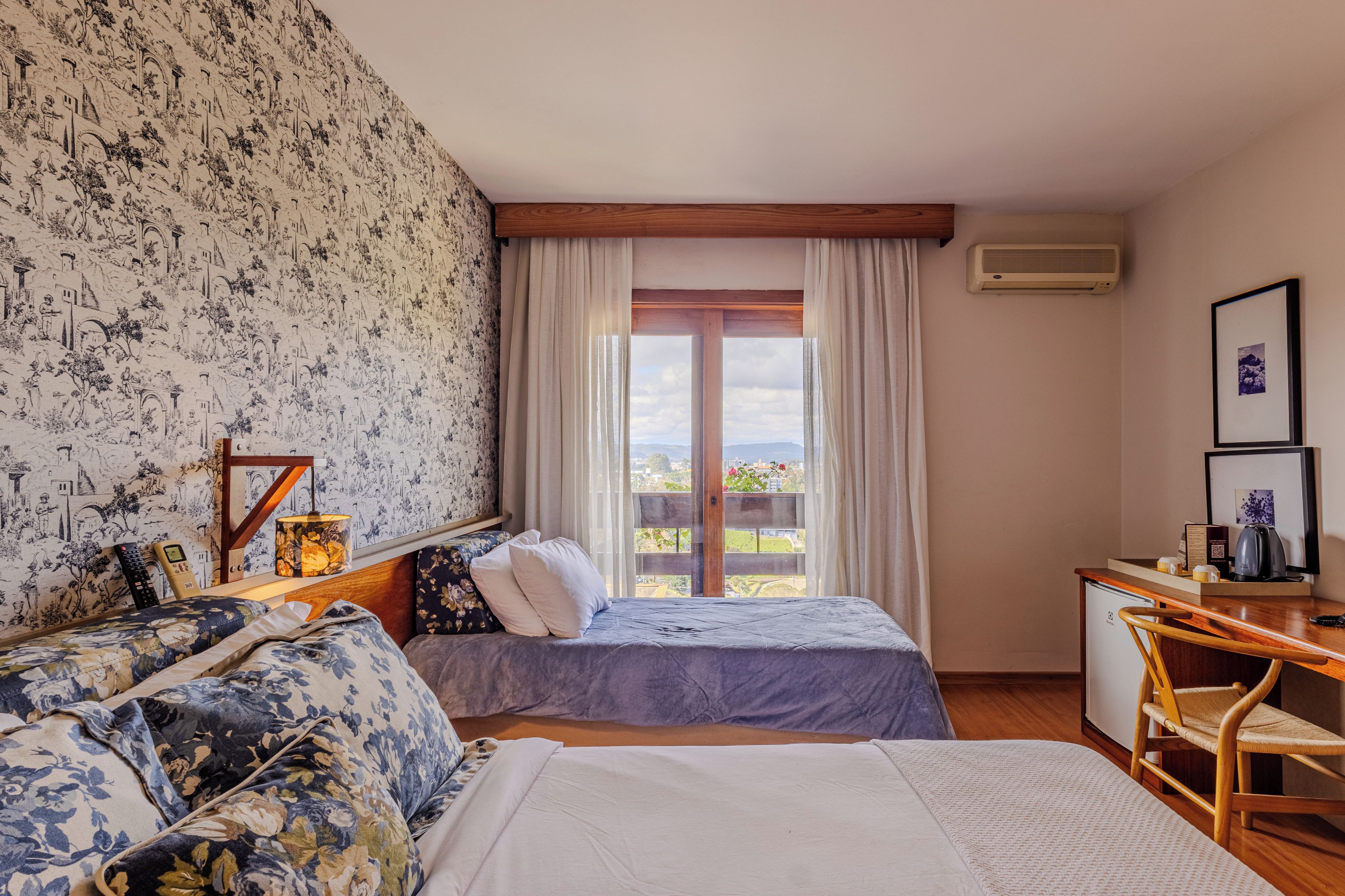 A hotel room with a bed, a desk, a chair, a window with curtains, and a wall with a patterned wallpaper.