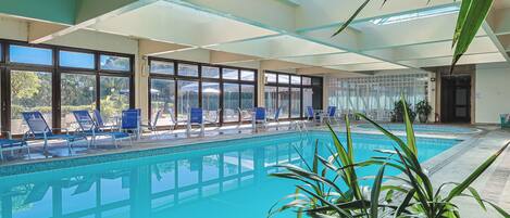 Una piscina interior con paredes de vidrio transparente, sillones reclinables azules y vistas al exterior.
