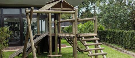 Eine hölzerne Spielkonstruktion mit Rutsche und Kletterleiter, eingebettet in einen grünen Garten mit einem Gebäude im Hintergrund.
