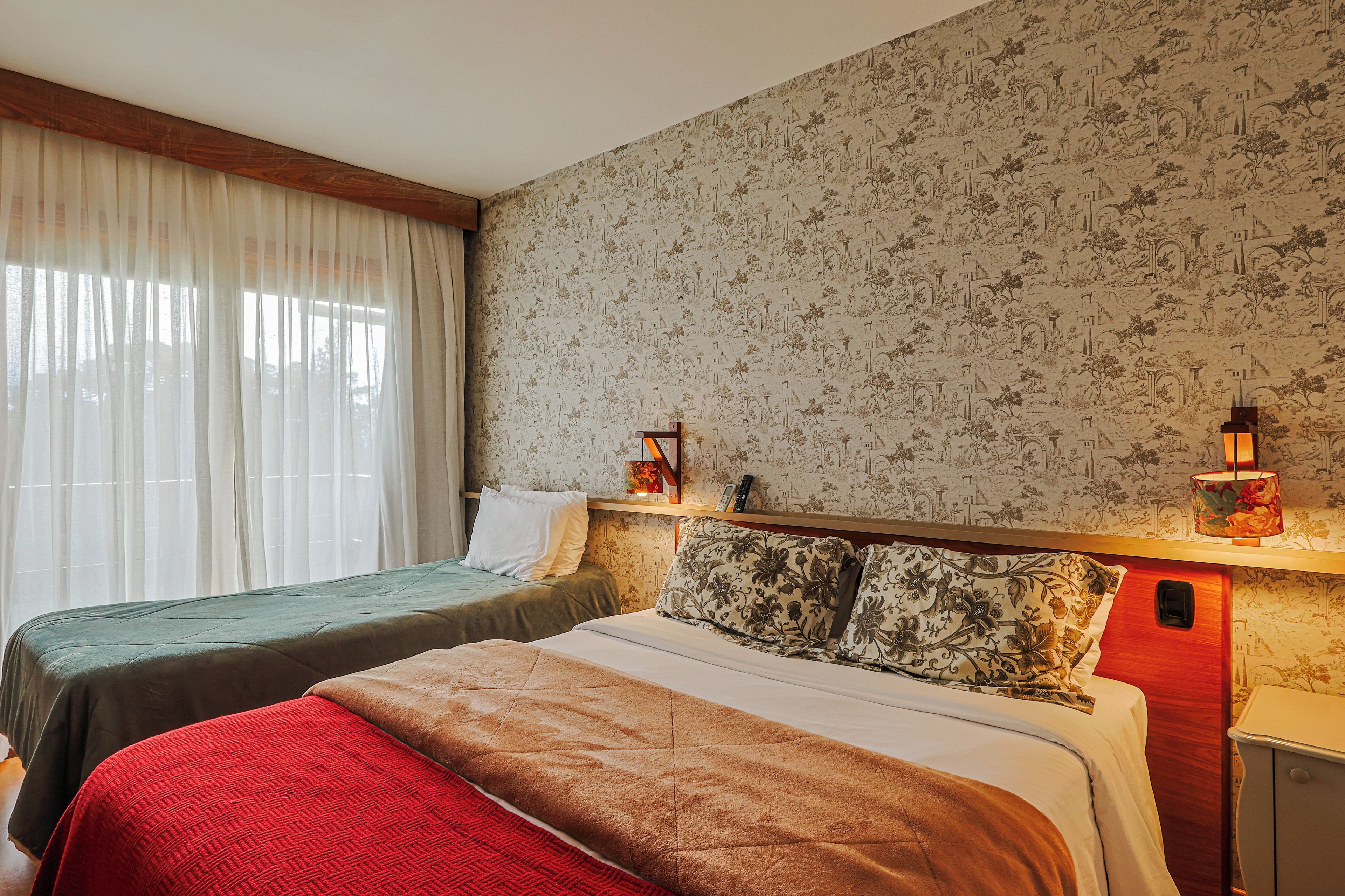 Ett hotellrum med två sängar, ett stort fönster med tunna, genomskinliga gardiner, en mönstrad tapet och sänglampor.