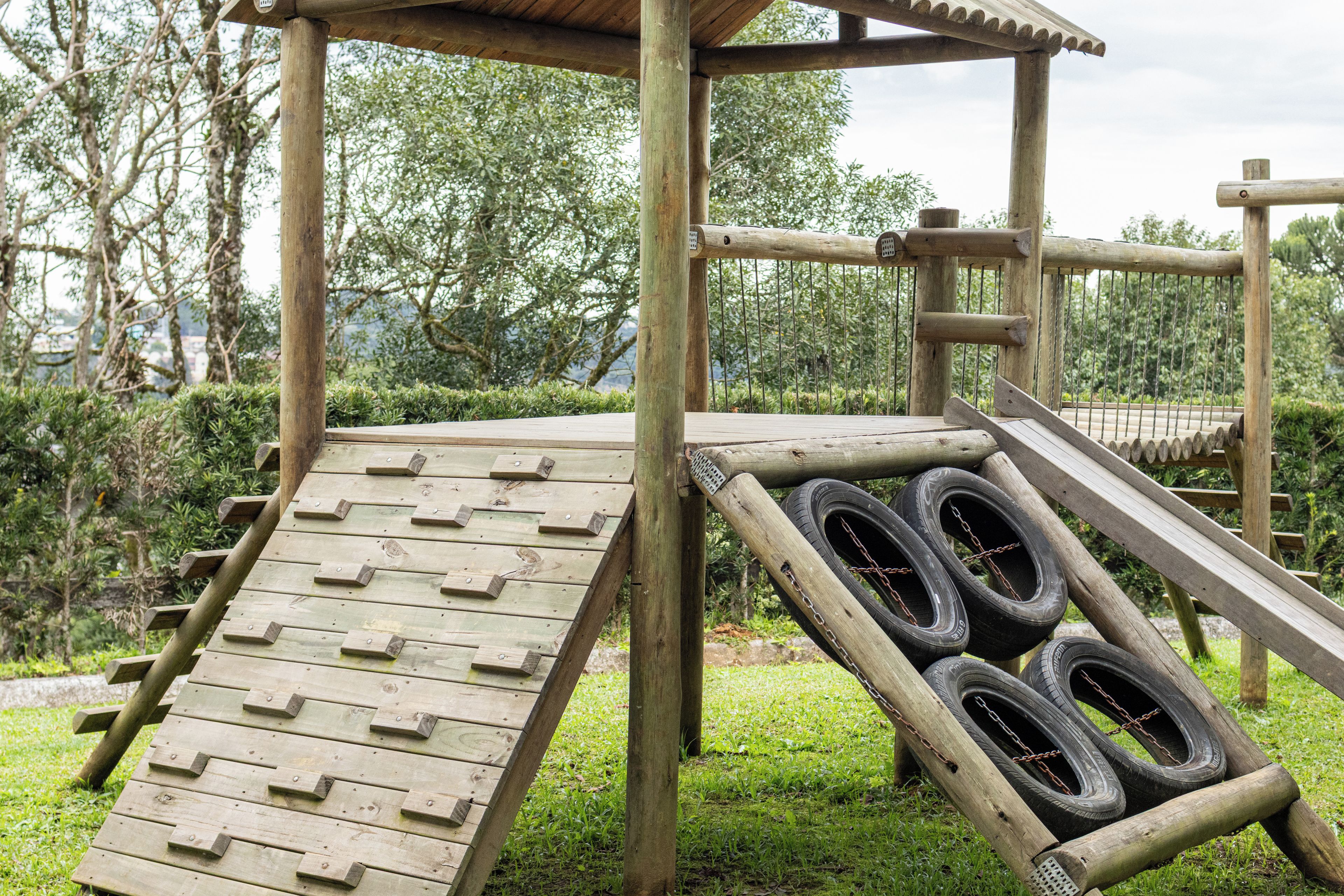 A wooden play structure with a slide and climbing area, surrounded by greenery.