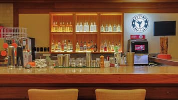 Uma área de bar com balcão, prateleiras cheias de garrafas de bebidas, uma máquina de café e uma tela de televisão.