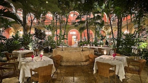 Un espace de restauration intérieur avec des tables disposées pour deux personnes, entouré d’une végétation luxuriante et agrémenté d’une structure centrale en pierre.