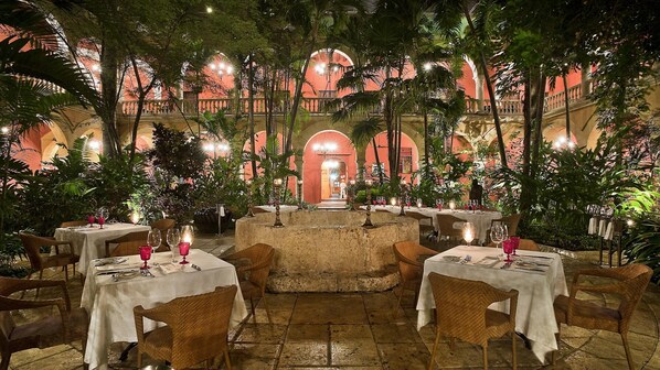 Un espacio de comedor al aire libre con mesas dispuestas para dos, rodeado de abundante vegetación y un suelo de piedra.
