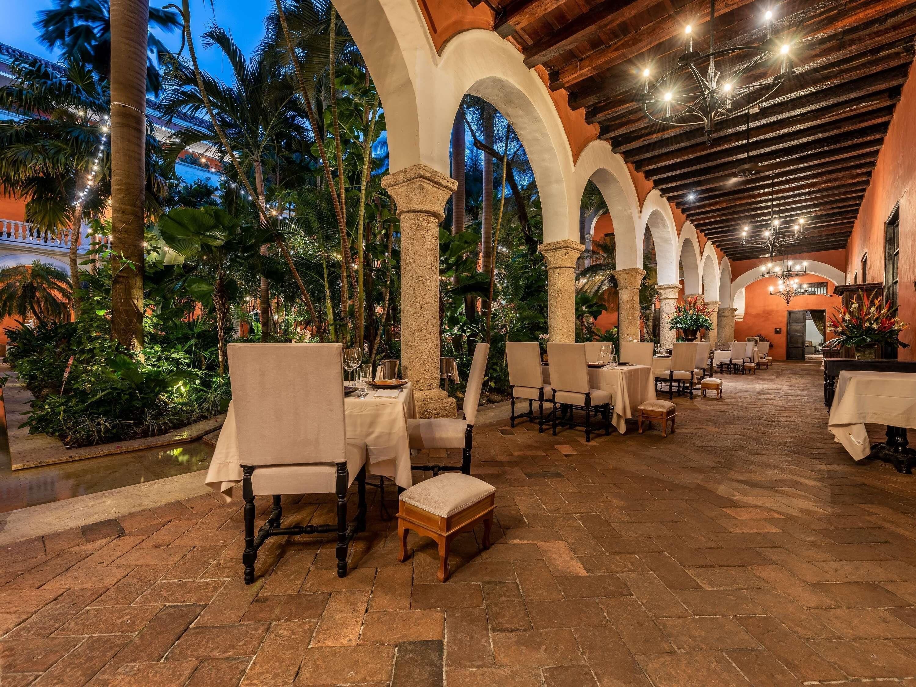 An indoor dining area with tables and chairs, surrounded by columns and arches, and a view of palm trees outside.