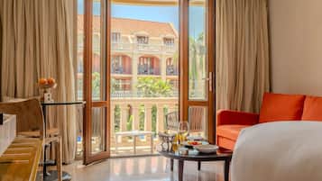 A hotel room with a bed, a table with a glass top, a sofa, and a balcony with a view of a building and palm trees.