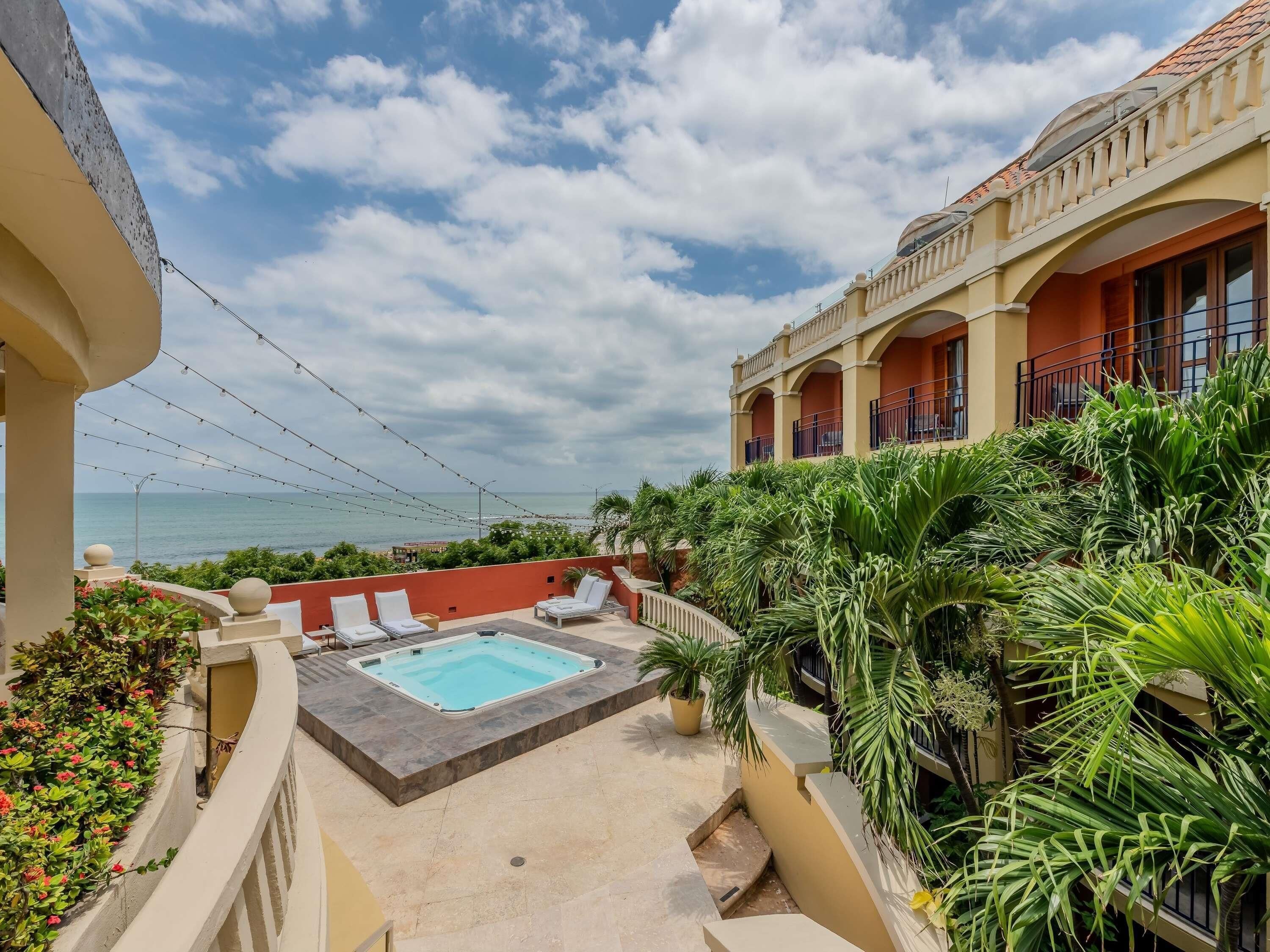 A rooftop pool with lounge chairs, surrounded by lush greenery and overlooking a beach.