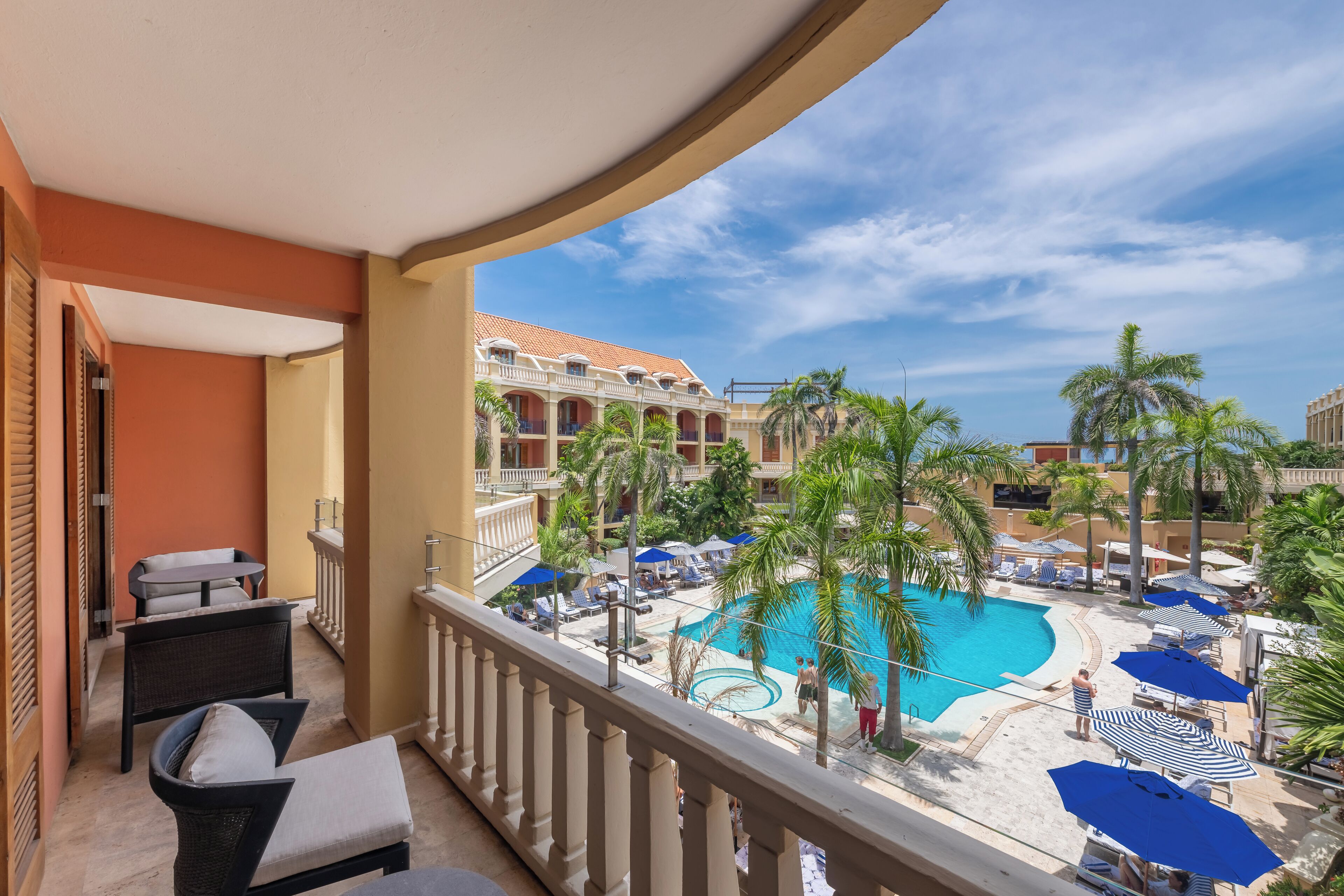 A balcony with wicker furniture, a pool area with umbrellas, and a view of a hotel building.