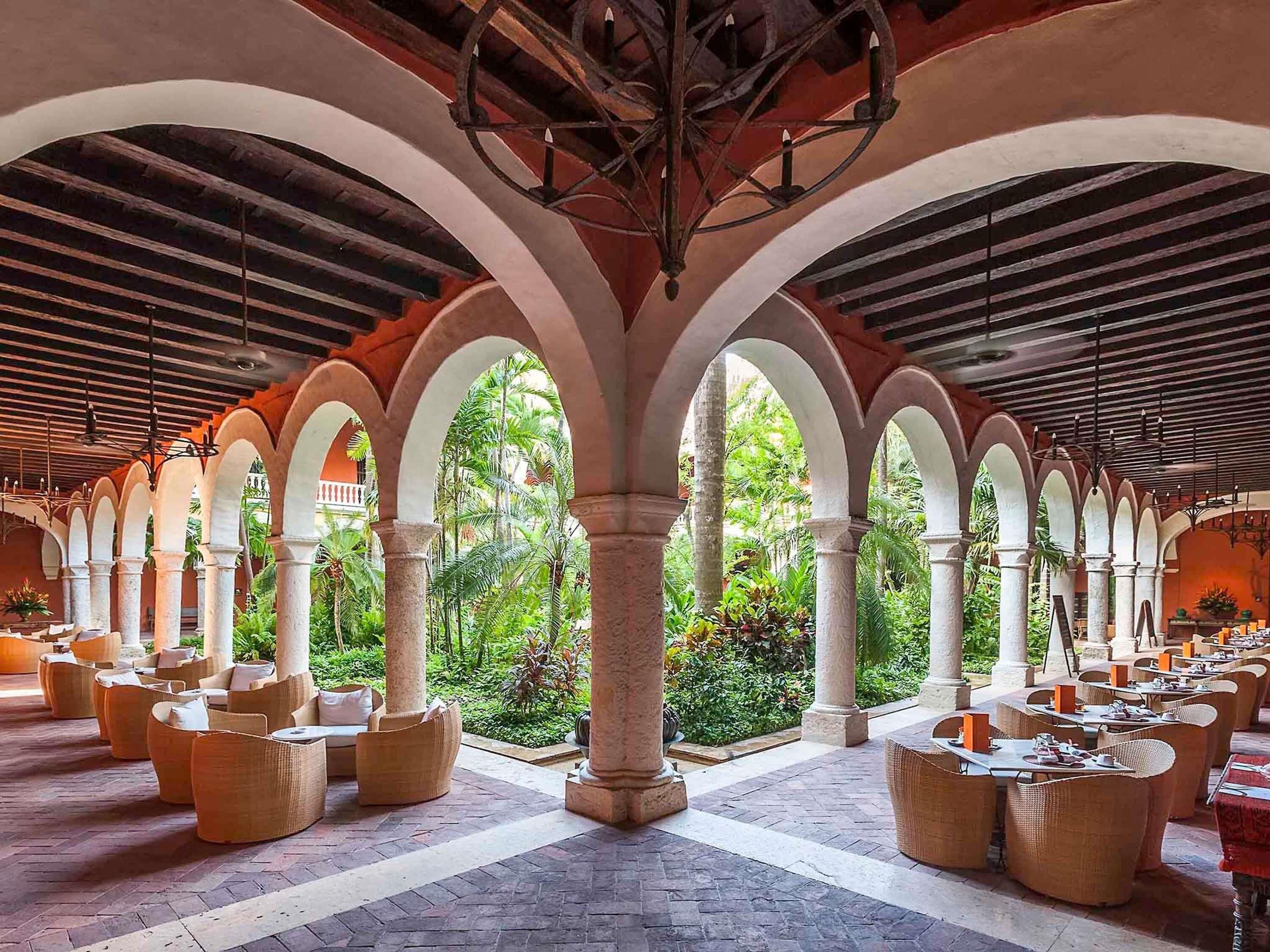 A covered outdoor seating area with columns and arches, surrounded by greenery.