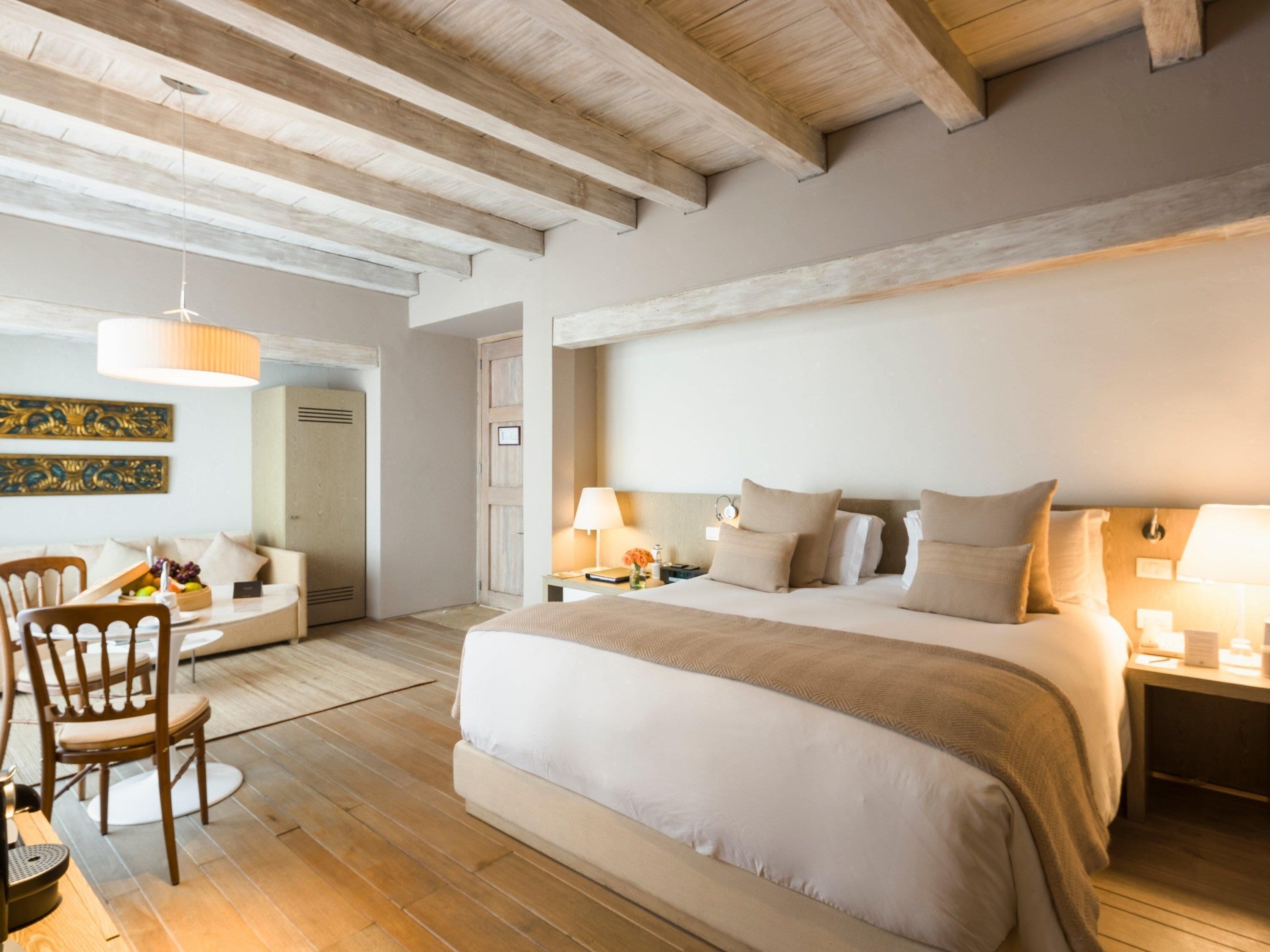A bedroom with a large bed, wooden beams on the ceiling, a desk with a chair, and a dining area with chairs and a table.