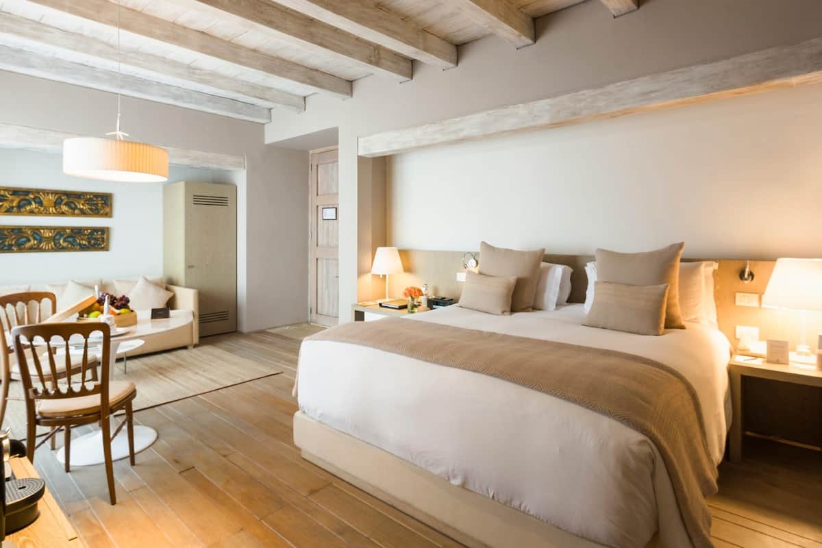 A bedroom with a large bed, wooden beams on the ceiling, a desk with a chair, and a dining area with chairs and a table.