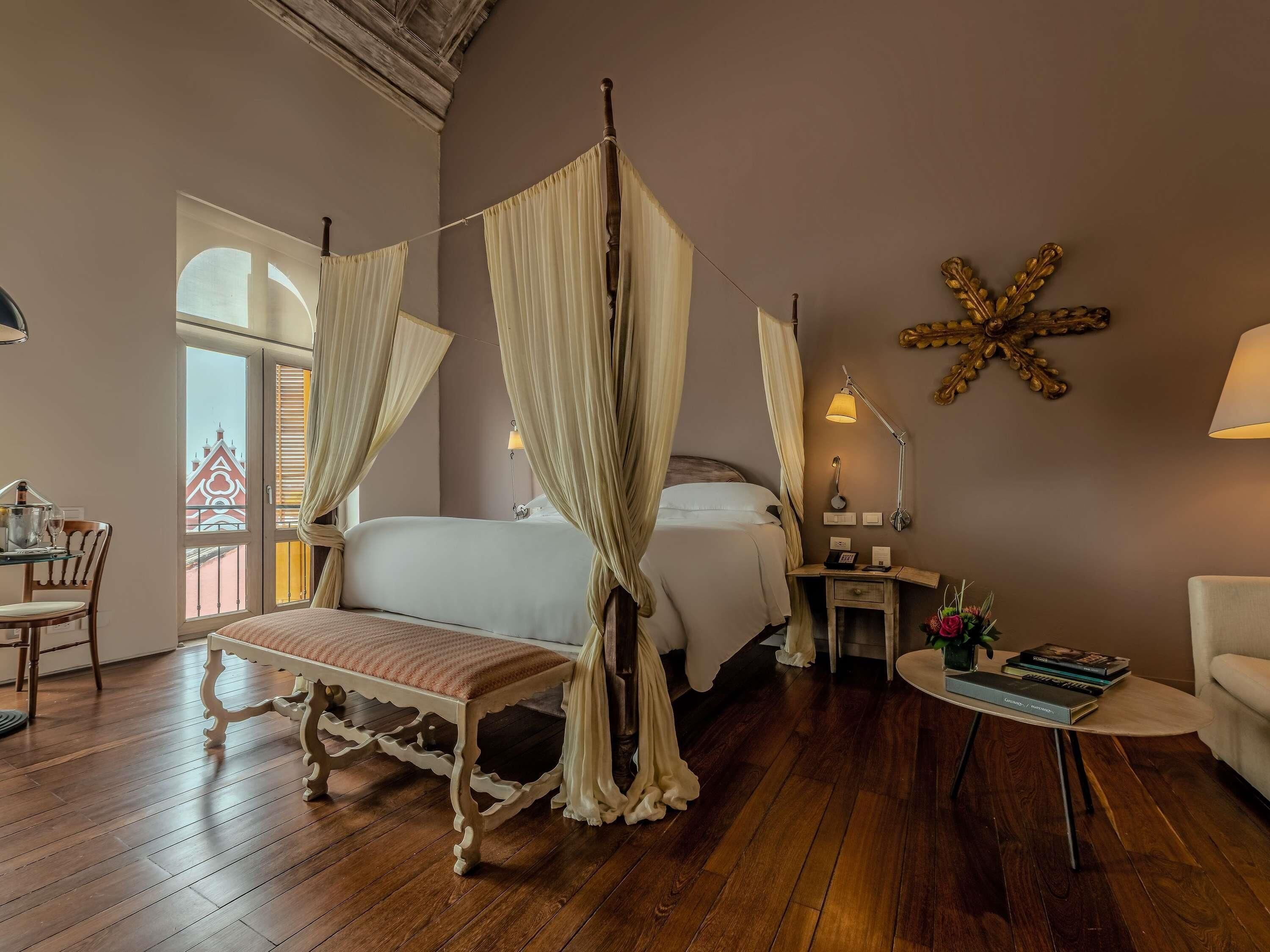 A room with a four-poster bed, a bedside table with a lamp, a small round table with books, and a chair.