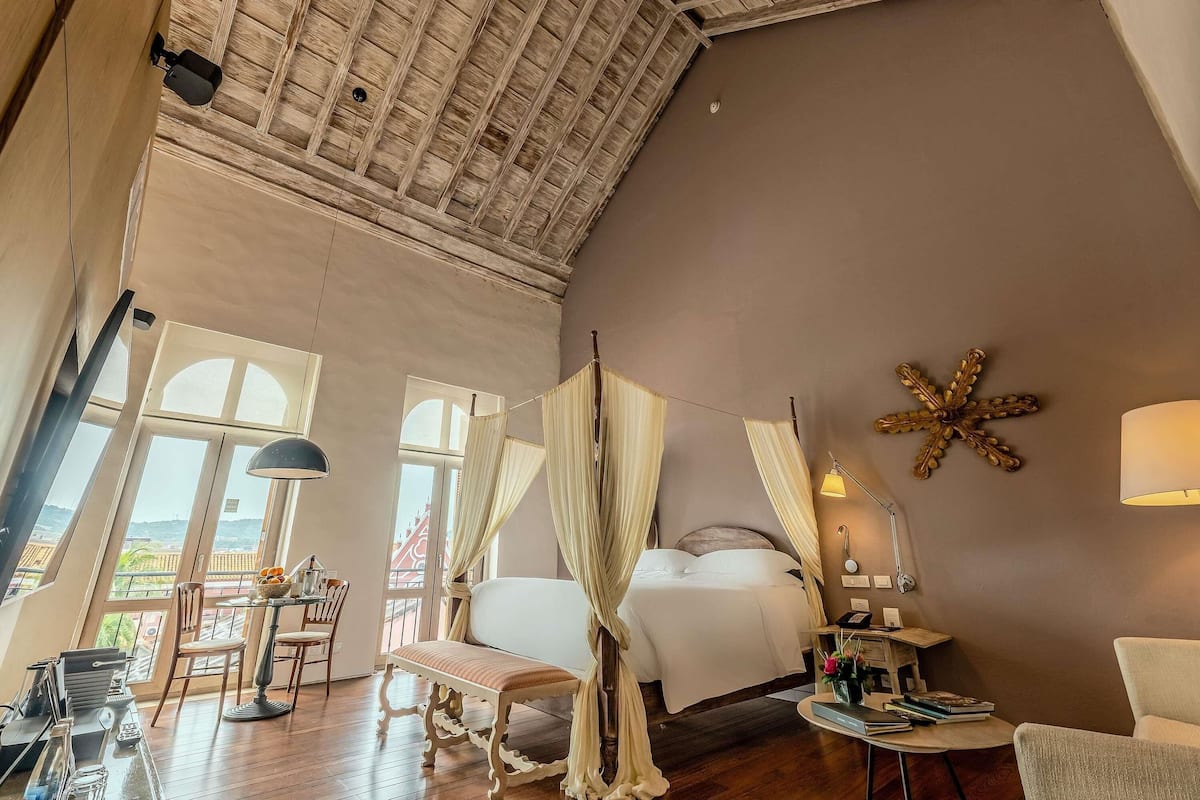 Una habitación amplia con techo alto, una cama con ropa blanca, un escritorio con lámpara y una silla.