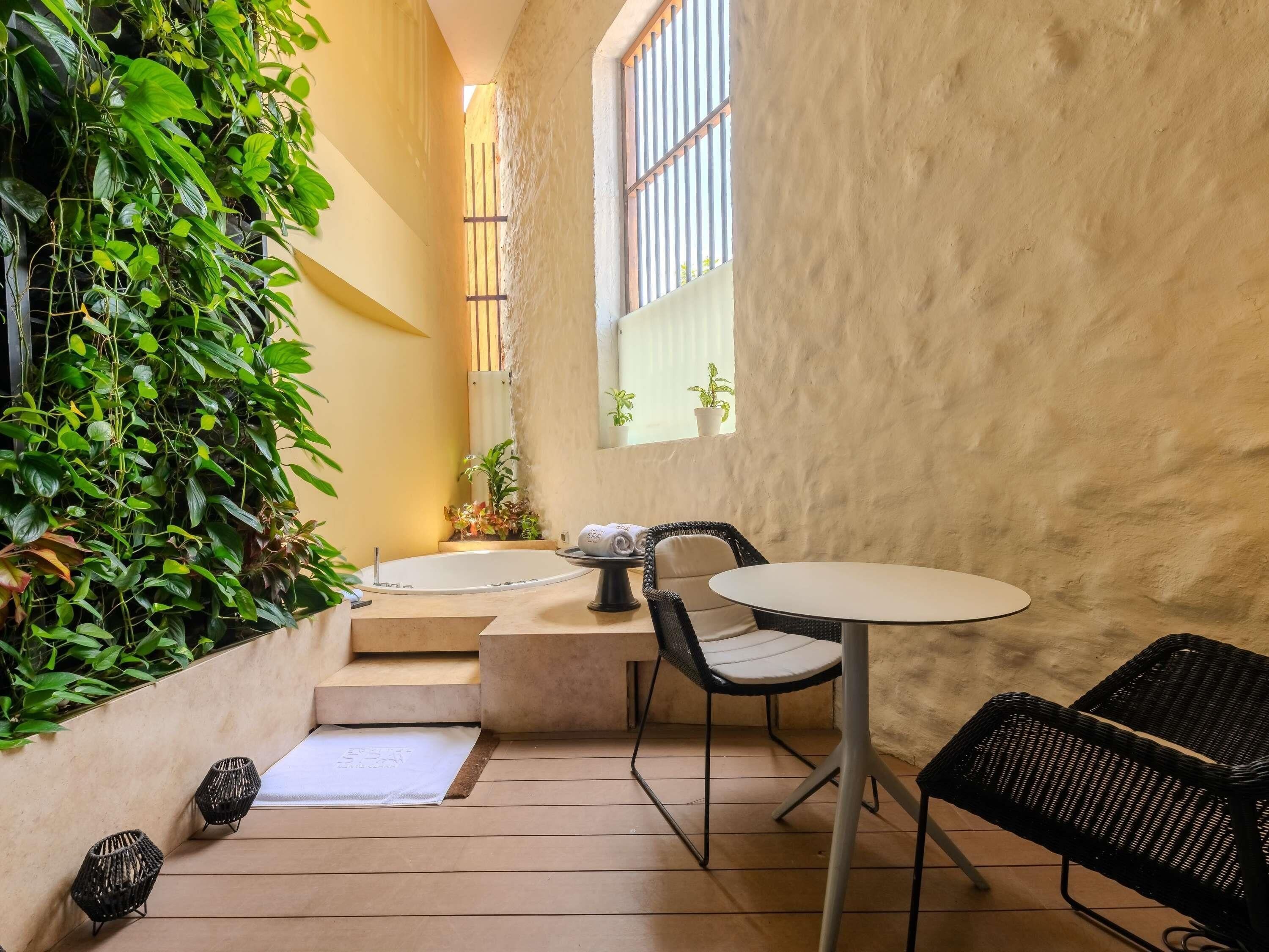 Un espacio exterior pequeño y acogedor con una mesa redonda y sillas, una bañera blanca y abundante vegetación.
