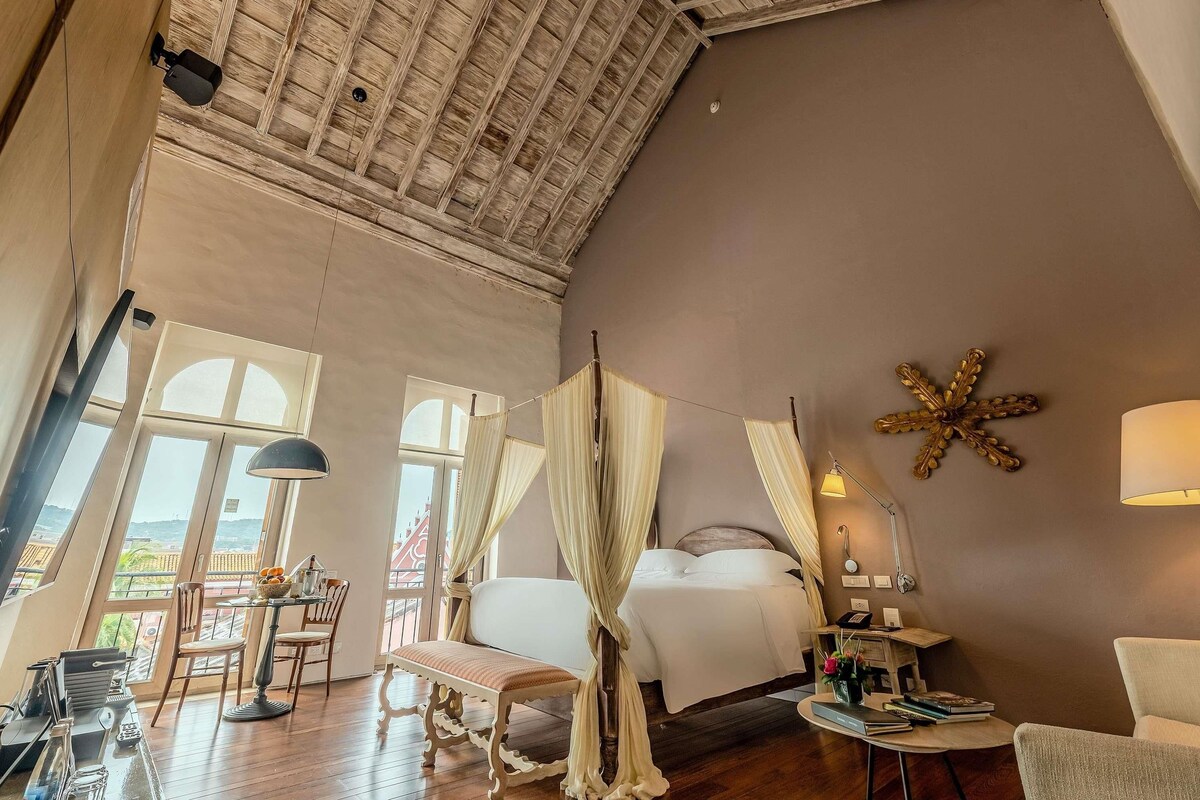 A spacious room with a high ceiling, a bed with white bedding, a desk with a lamp, and a chair.