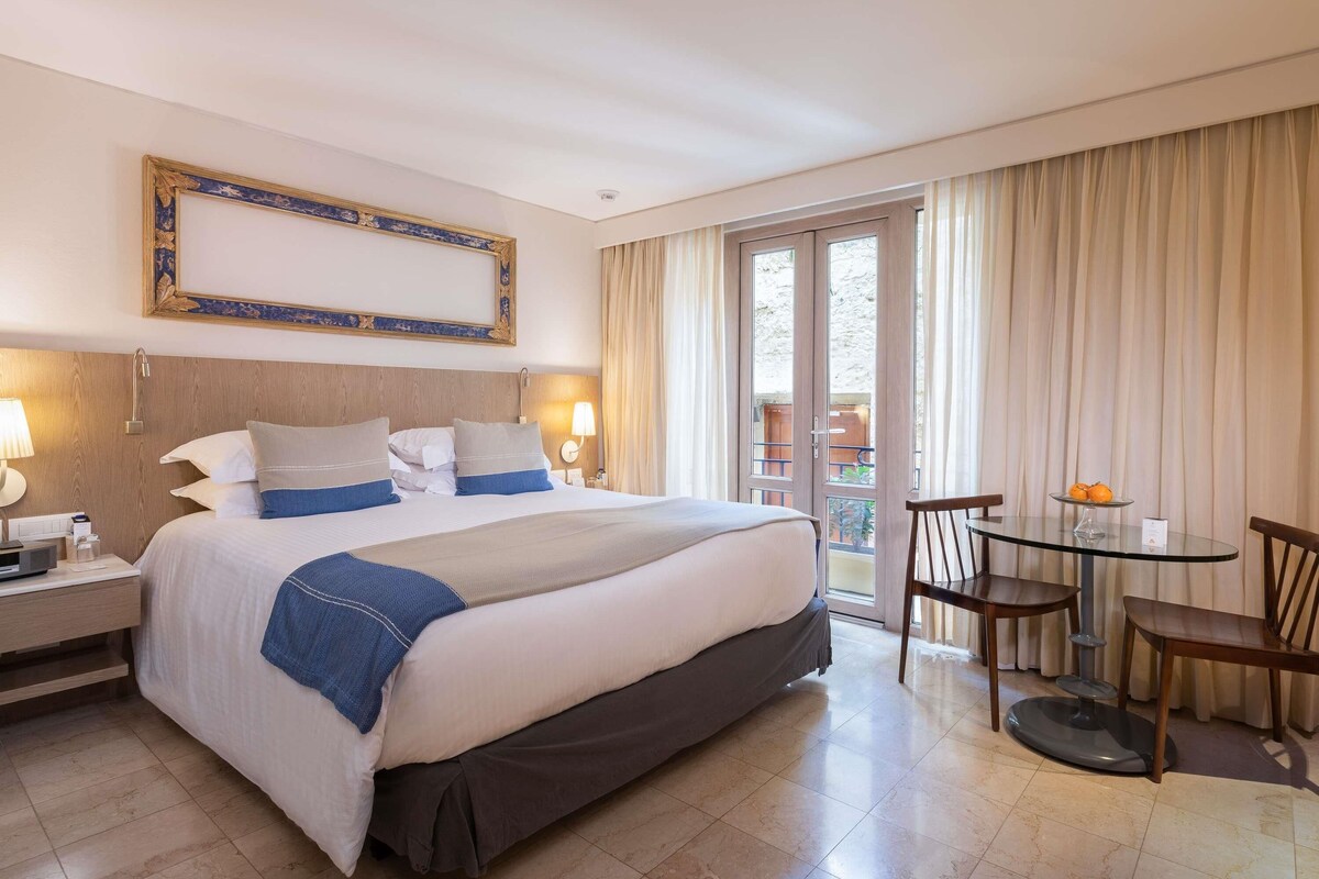A hotel room with a large bed, bedside lamps, a table with chairs, and a view through the glass door.