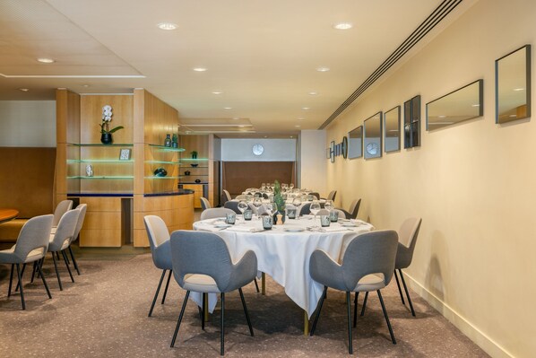 Un coin repas avec une table ronde dressée pour six personnes, entourée de chaises à revêtement gris. On y trouve des étagères en verre ornées d’objets décoratifs et un comptoir en bois avec une horloge au mur.