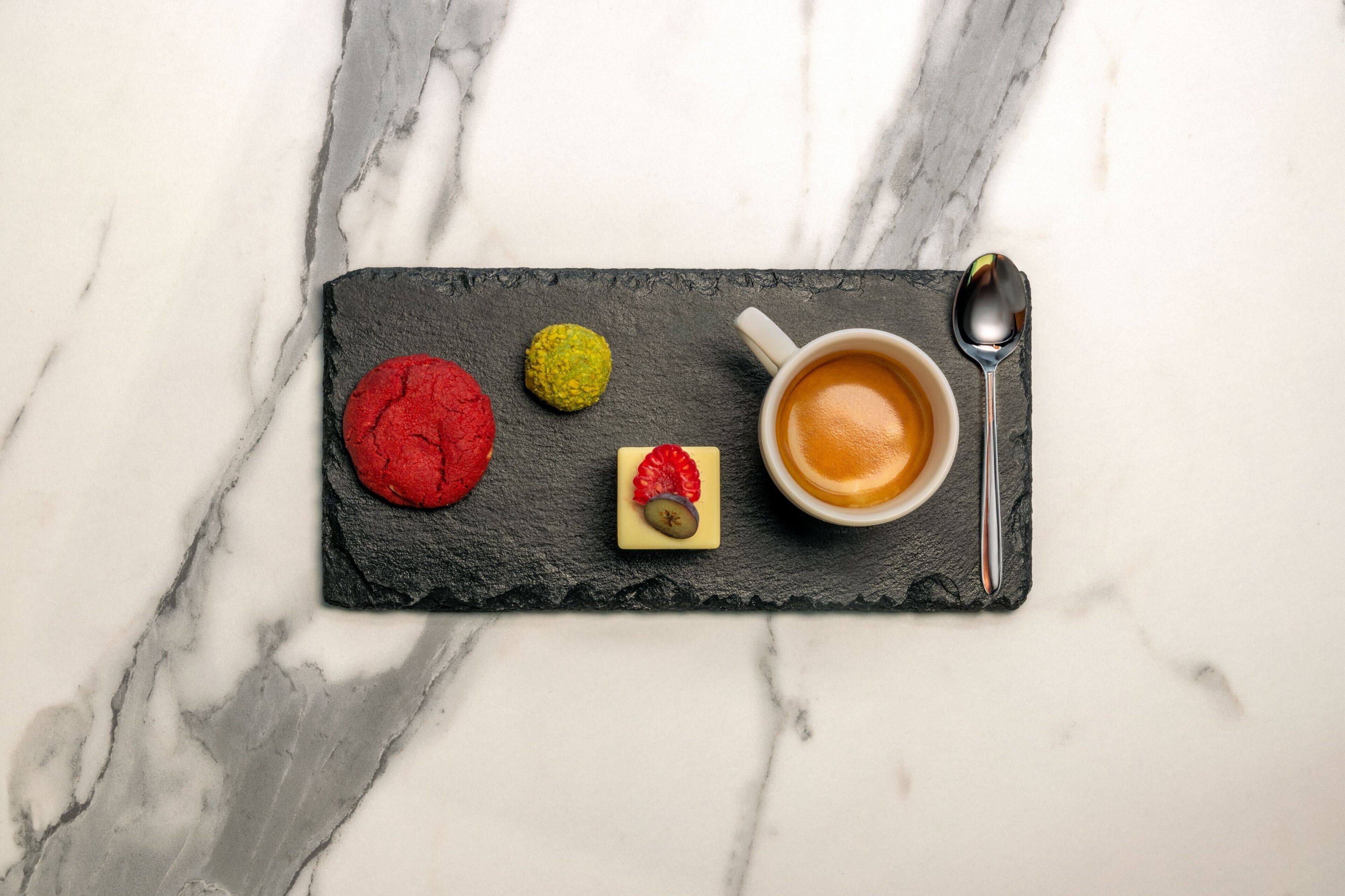 A slate serving board with a cup of coffee, a spoon, and three cookies on a marble surface.