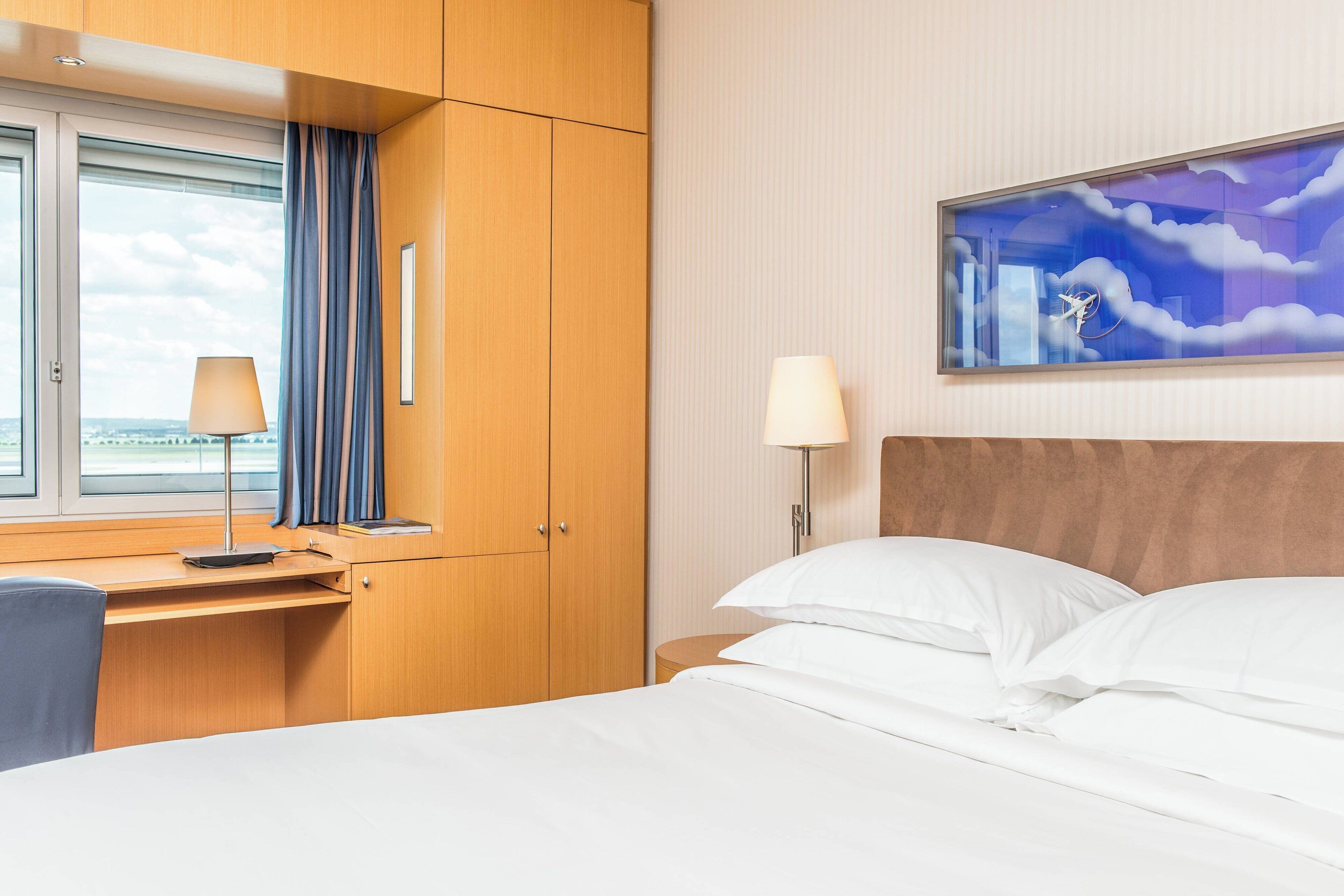 Une chambre d’hôtel avec un lit, un bureau, une chaise, une lampe et une fenêtre avec des rideaux.