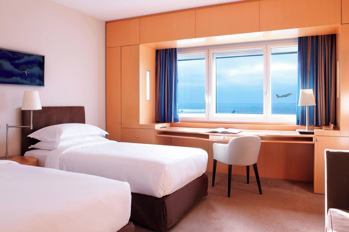 Habitación de hotel con dos camas, un escritorio, una silla y un ventanal con vistas al mar y un avión.