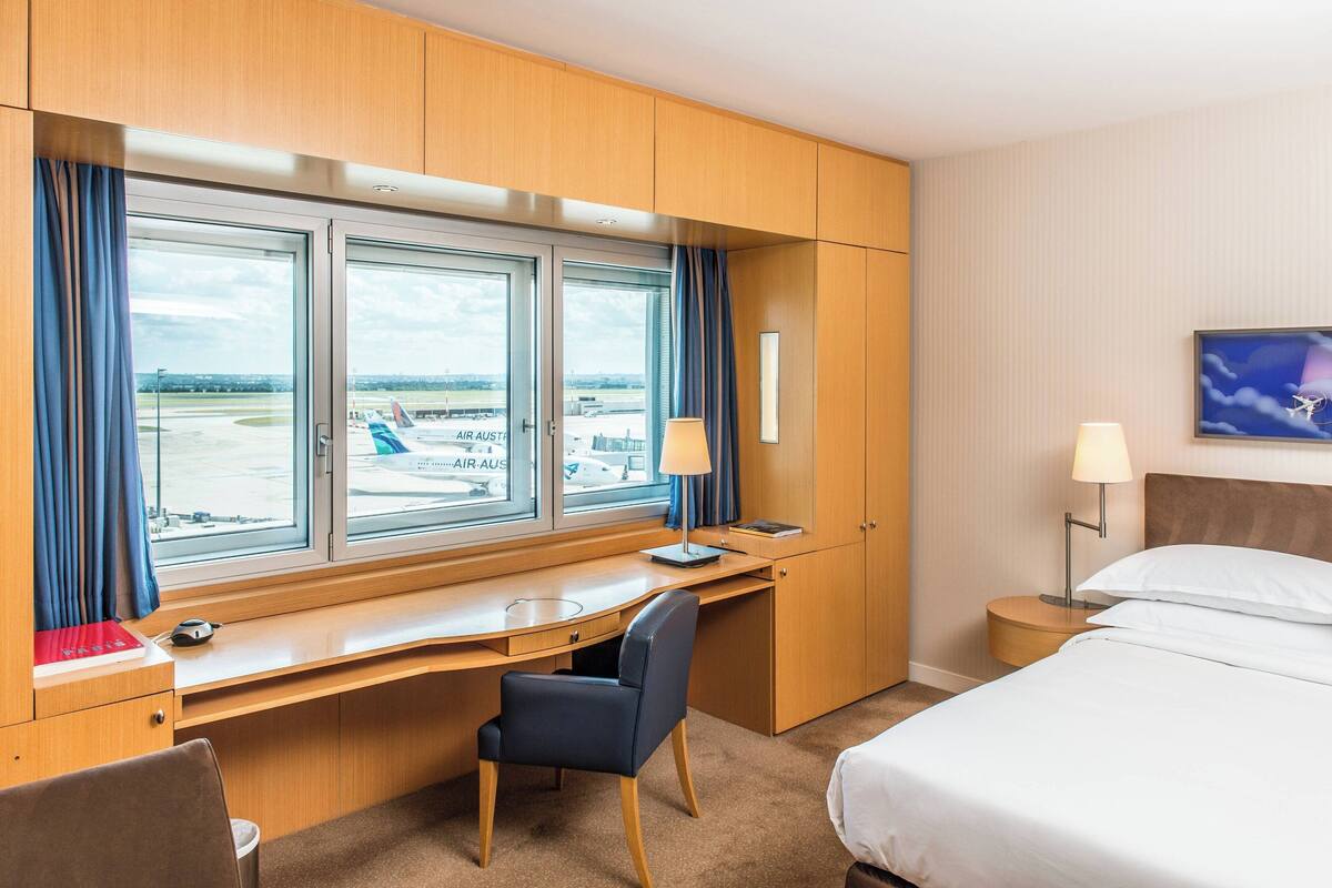 Habitación de hotel con escritorio, silla, cama y vistas al aeropuerto.