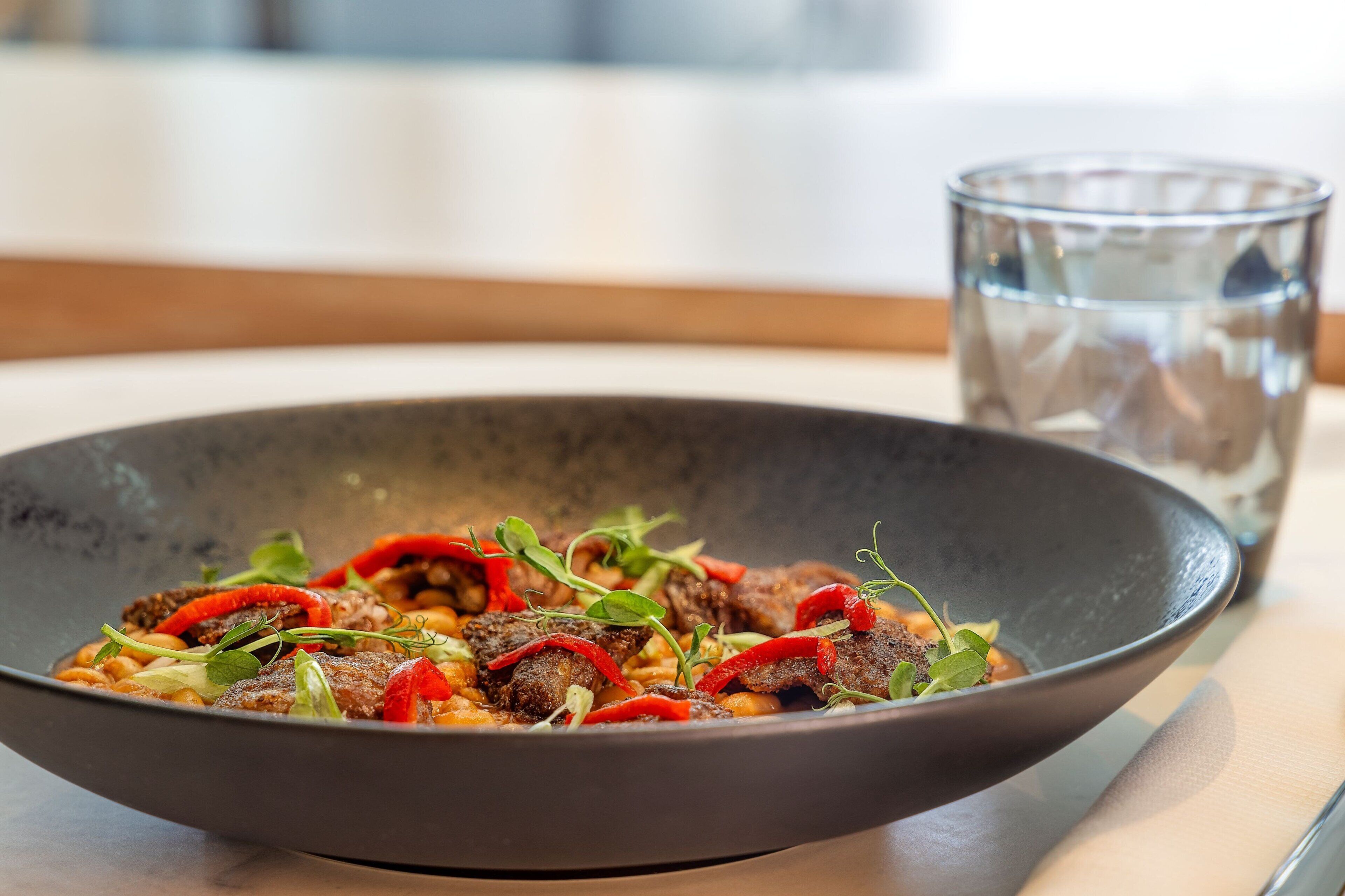 A bowl of food with red peppers and greens, a glass of water, and a wooden table.