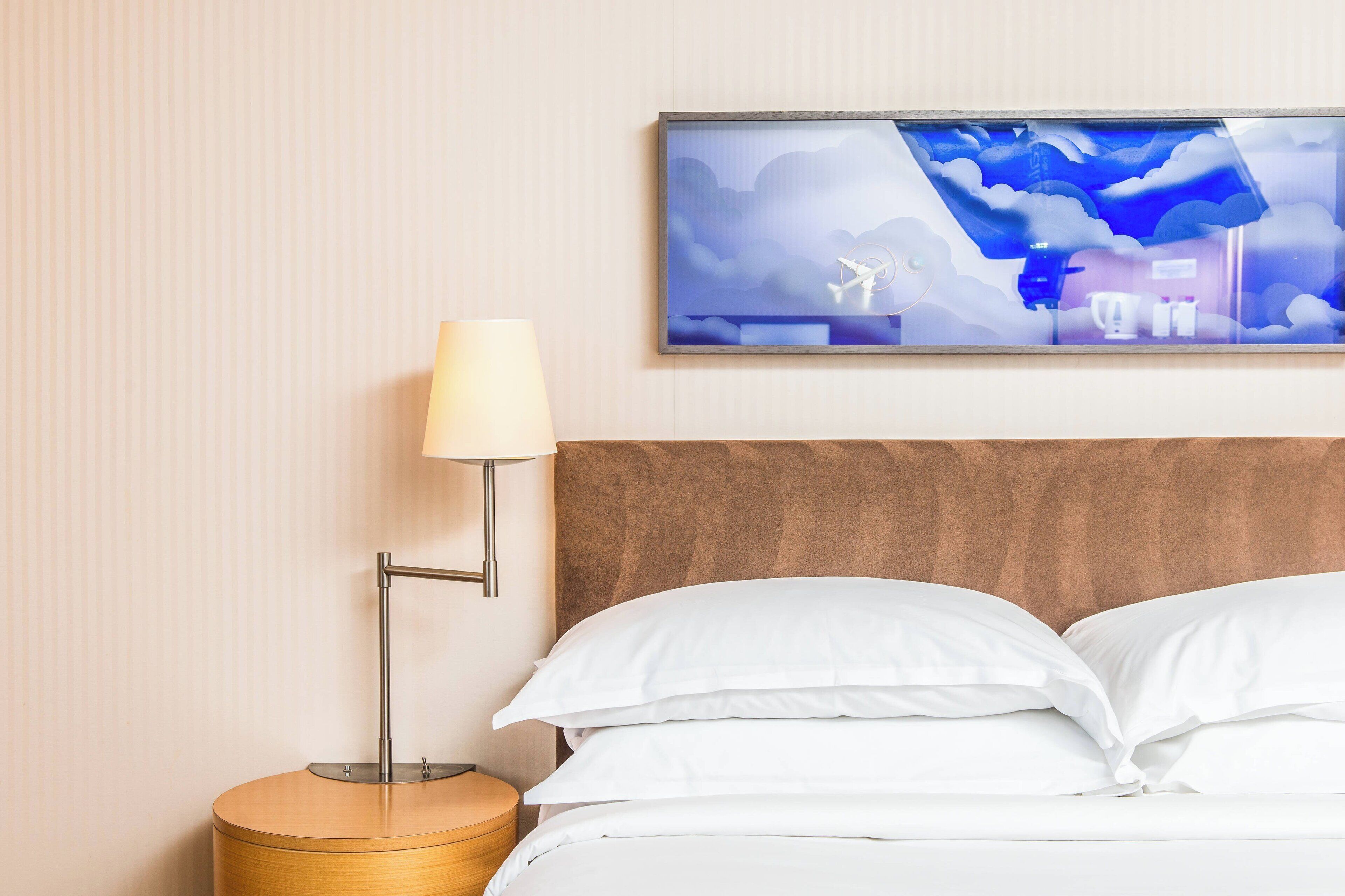 A neatly made bed with white linens, a wooden bedside table with a lamp, and a wall-mounted artwork depicting a stylized landscape.