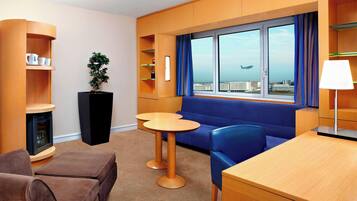A modern hotel room with a blue sofa, wooden tables, a small potted plant, and a view of the city through large windows.