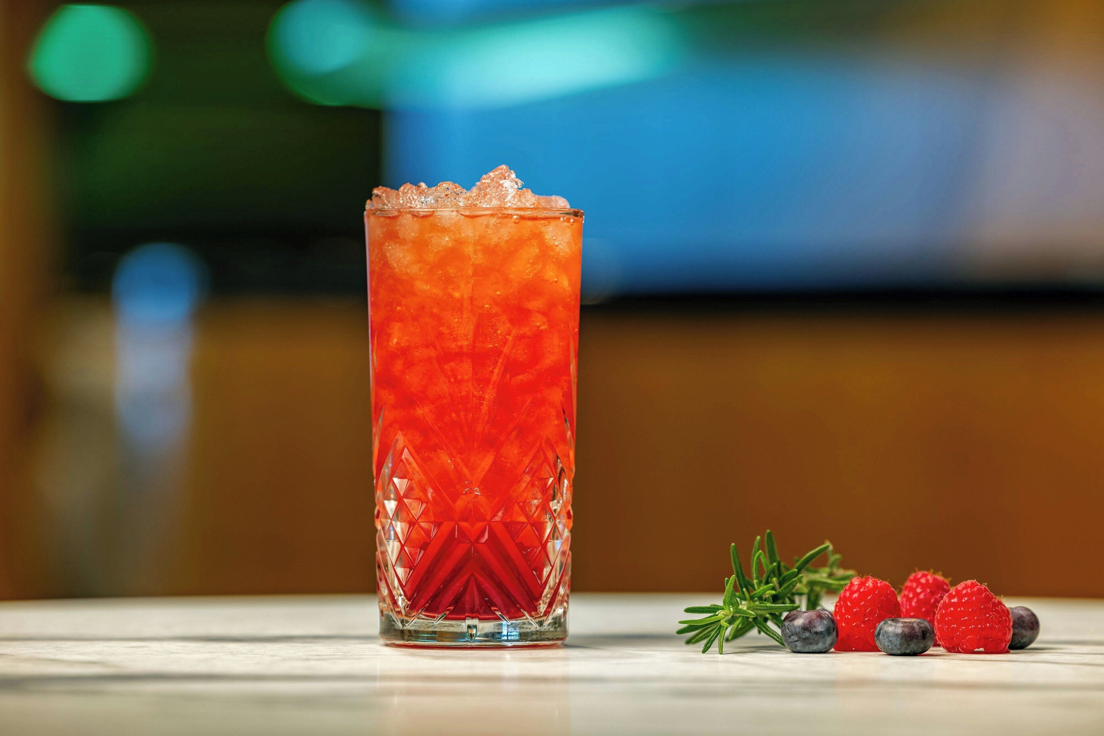 A tall glass with a textured pattern filled with a red beverage, ice cubes, and surrounded by fresh berries and a sprig of rosemary on a reflective surface.