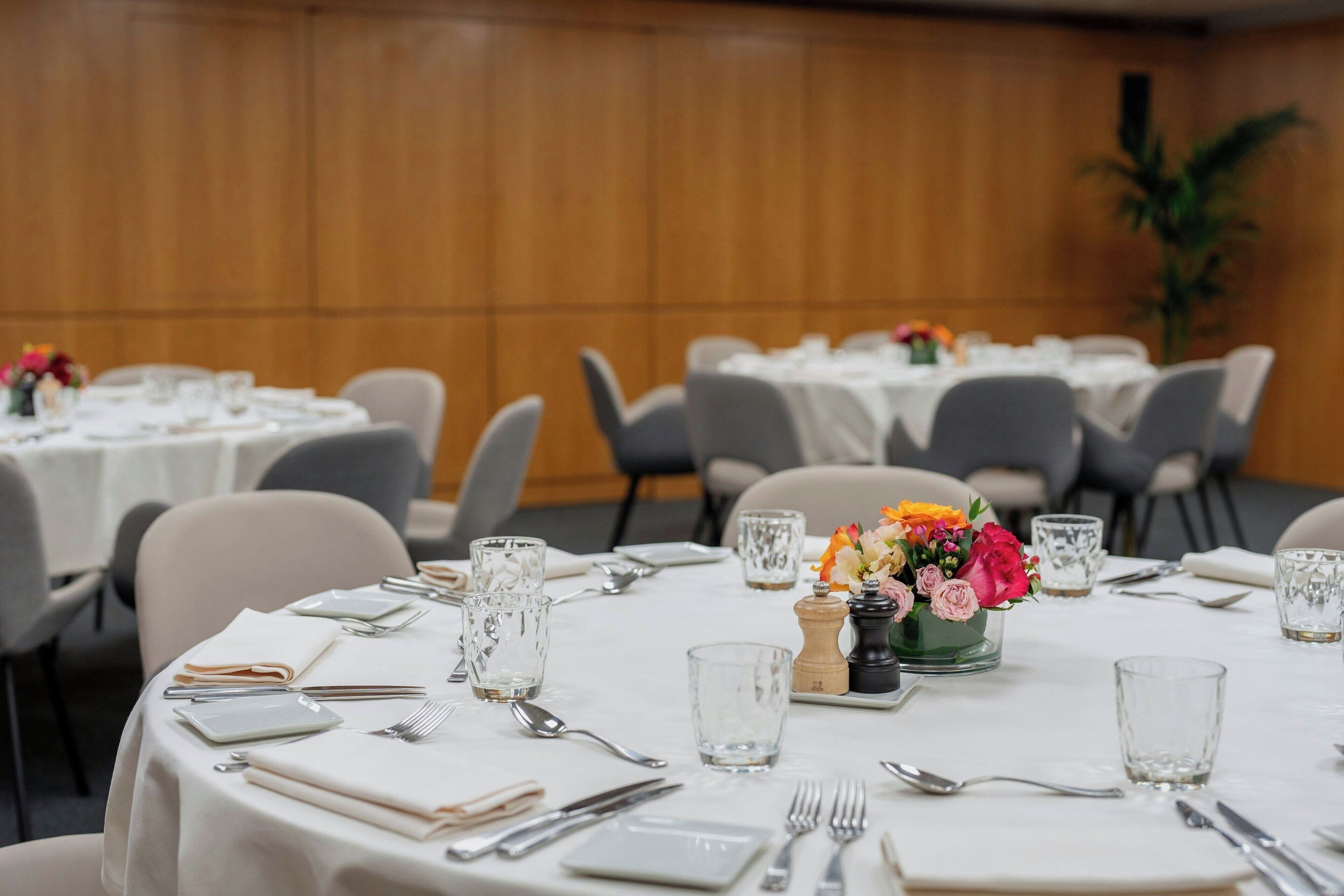 A conference room with round tables set for a meeting. Each table has a white tablecloth, plates, cutlery, and glasses. There are floral centerpieces on some tables.