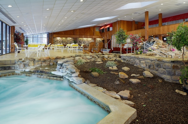 An indoor pool area with a stone waterfall, wooden seating, and a view of the outside through large windows.