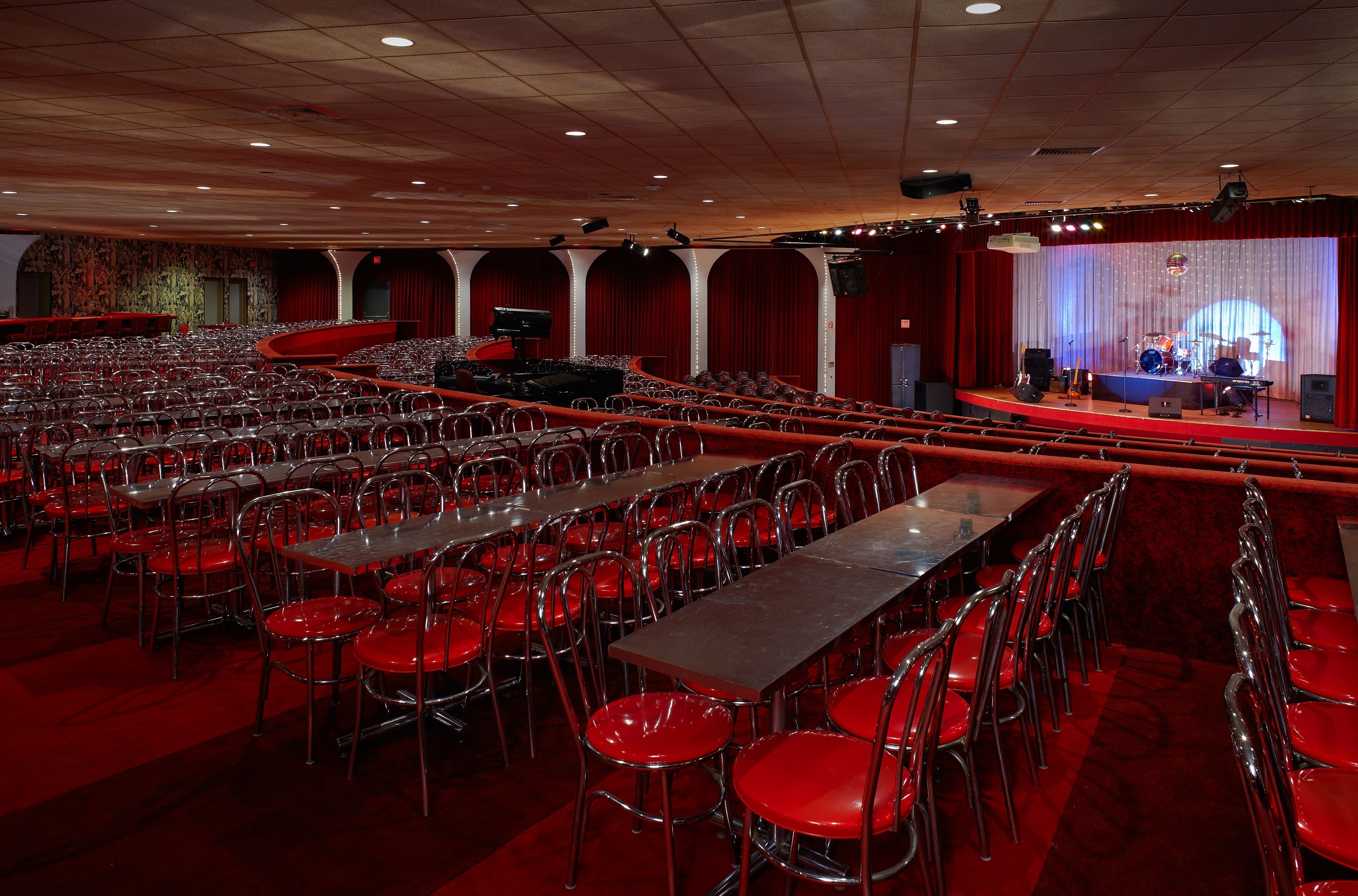 A large auditorium with rows of red chairs and tables, a stage with musical instruments, and a red carpeted floor.