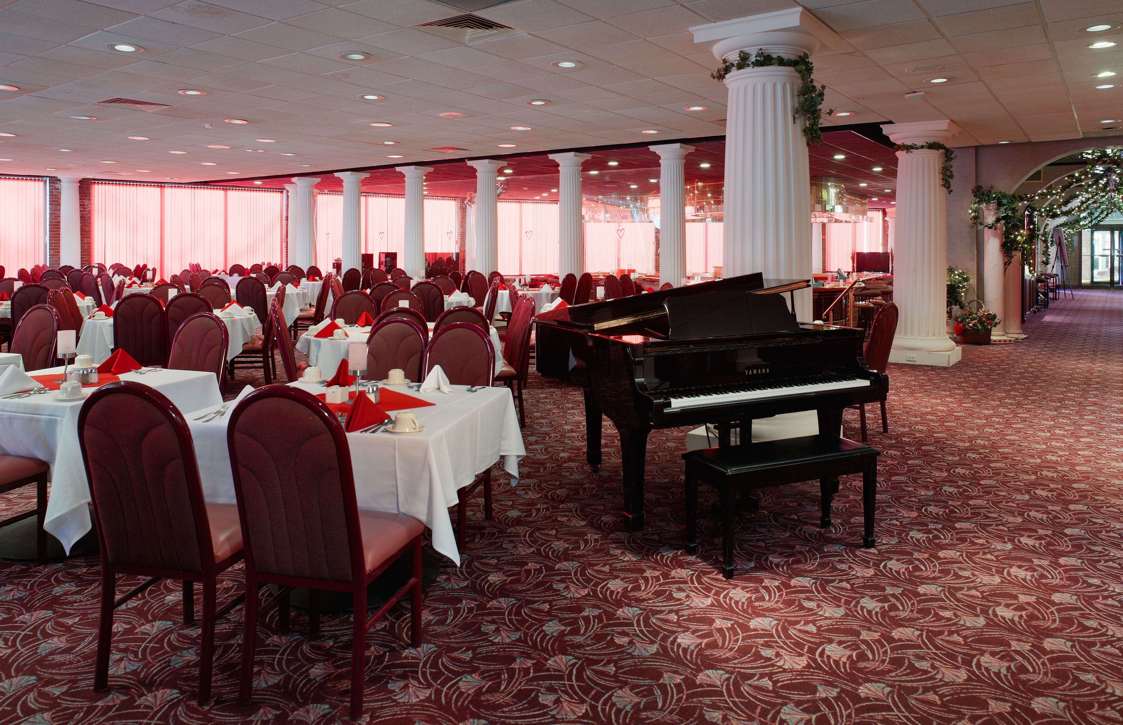 A banquet hall with round tables, red chairs, a grand piano, and white columns.