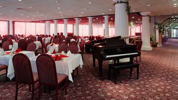 A banquet hall with round tables, red chairs, a grand piano, and white columns.