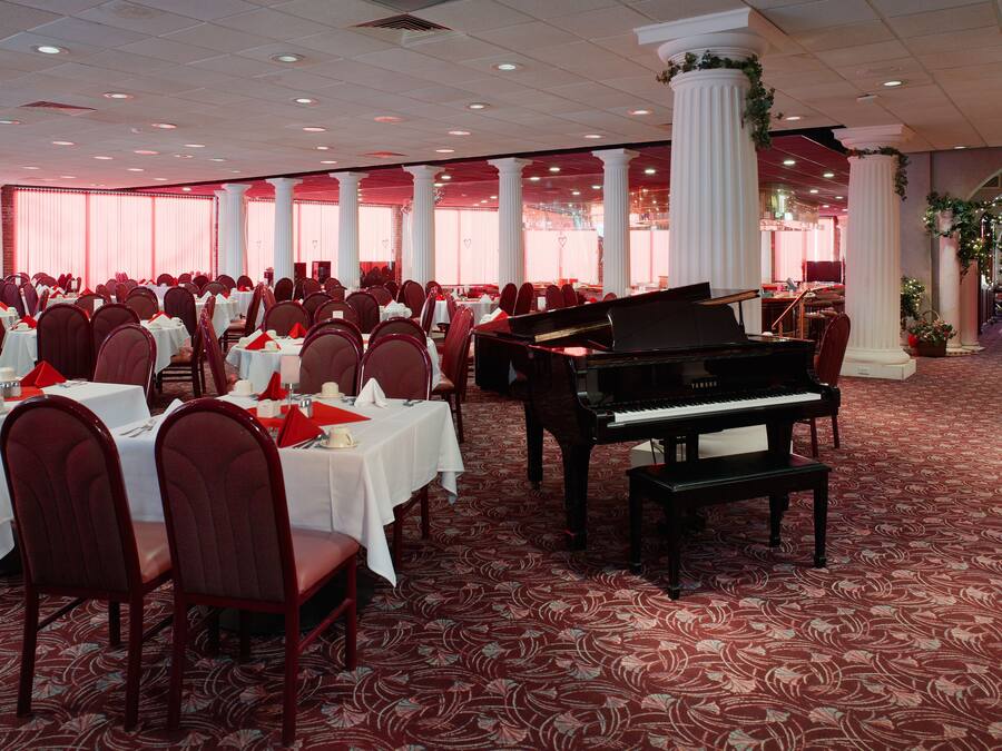 A banquet hall with round tables, red chairs, a grand piano, and white columns.