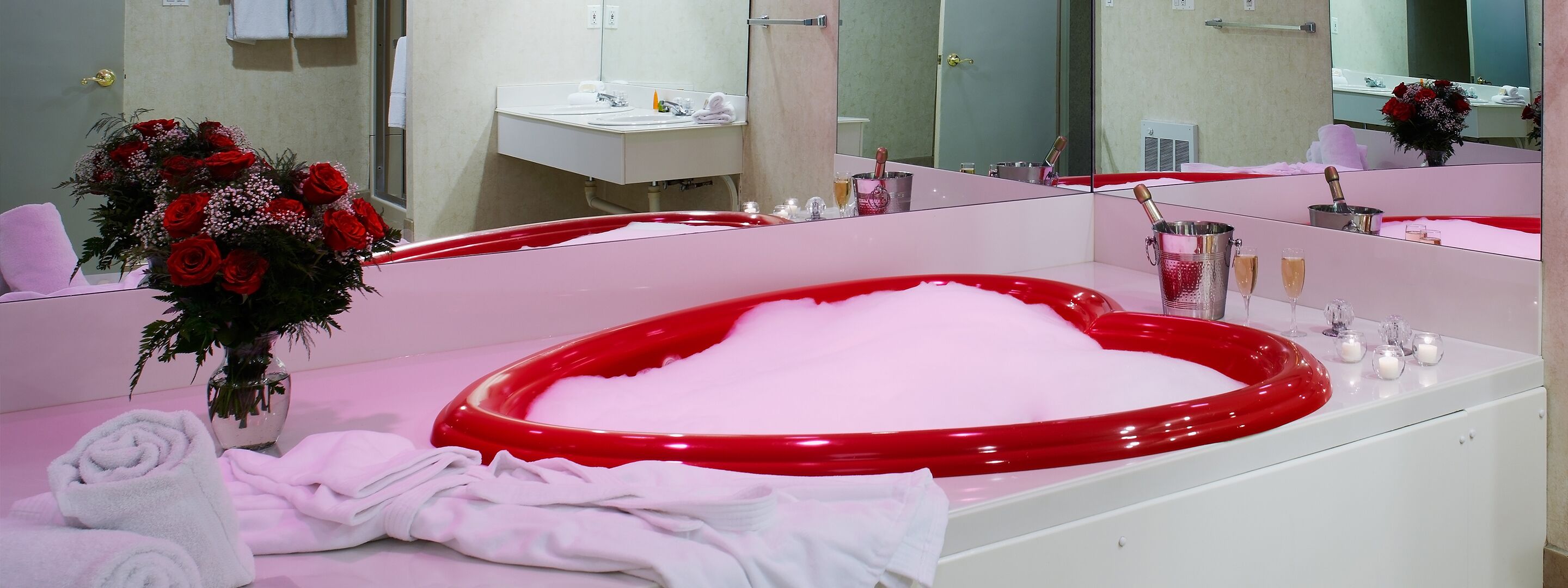 A bathroom with a red bathtub, white towels, a vase of red roses, and a mirror reflecting the room.