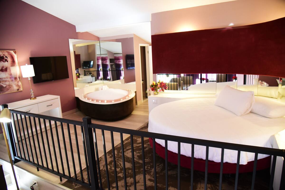 Une chambre d’hôtel avec un grand lit, une baignoire ronde, une coiffeuse avec un miroir et un téléviseur fixé au mur.